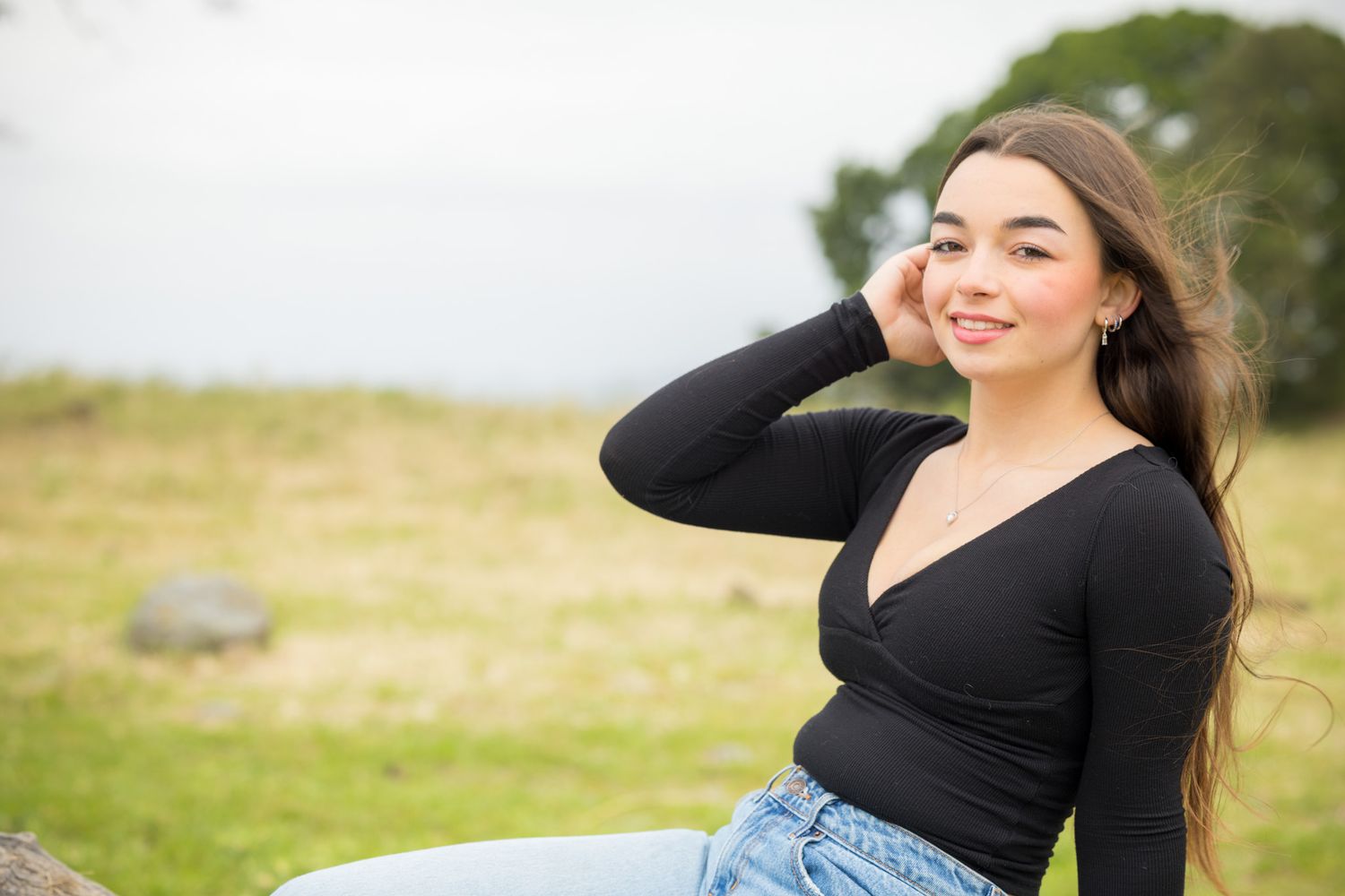 Young woman in black top and jeans sitting outdoors in a green field, smiling with hand near face.