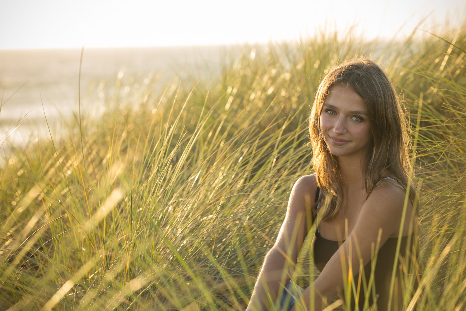 Young woman sitting among tall beach grass dunes at golden hour, smiling softly with ocean in background.