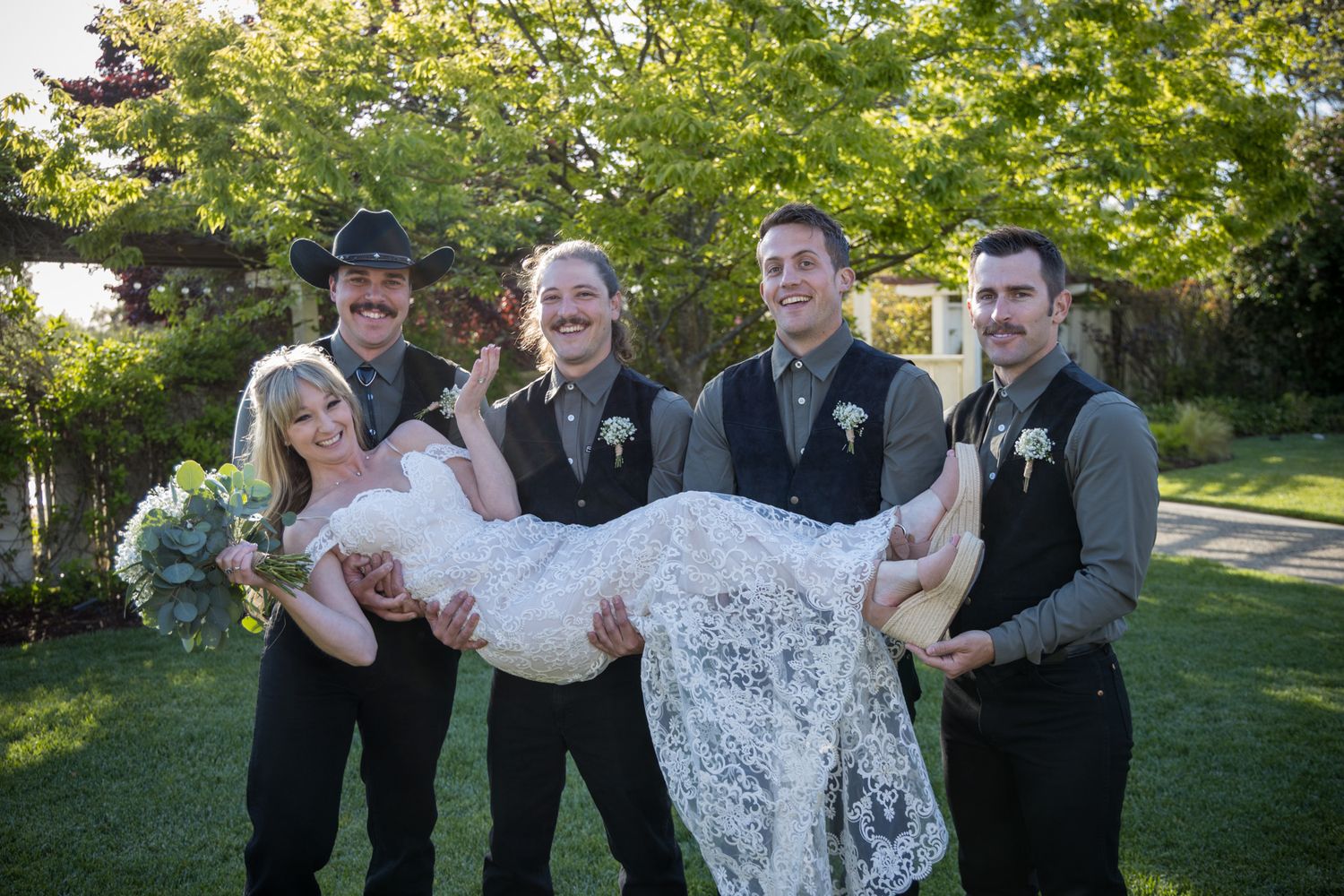 A group in formal wedding attire stands together in a garden setting while carrying someone in a white lace dress.