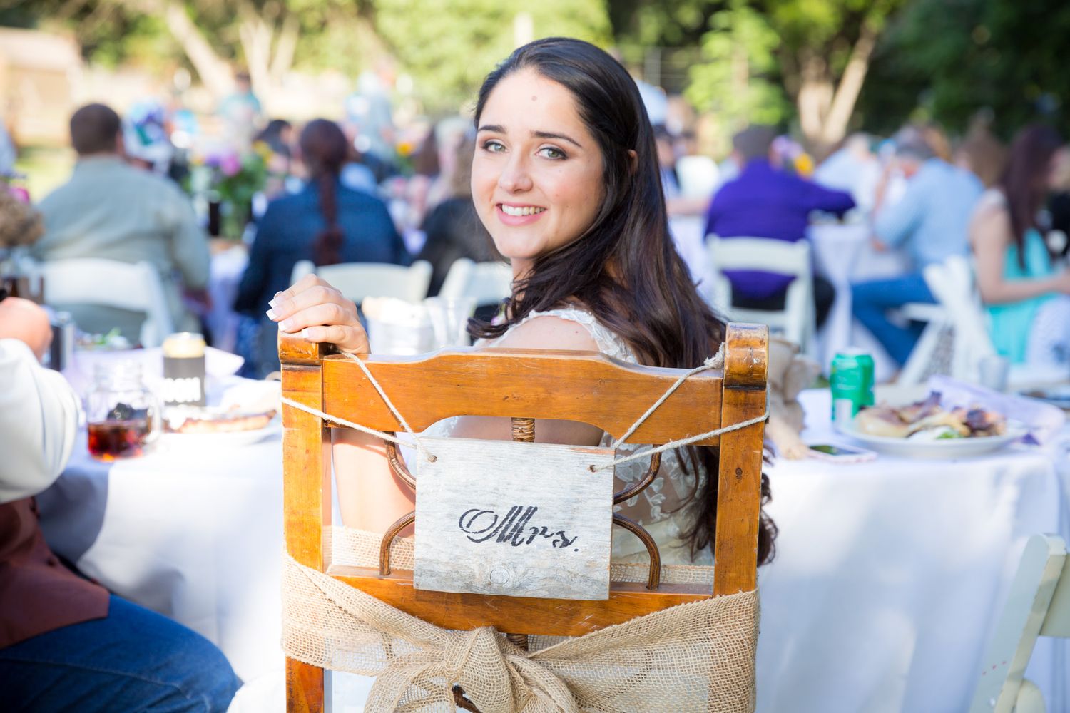 Someone sits smiling at an outdoor event with a wooden chair designation marked 