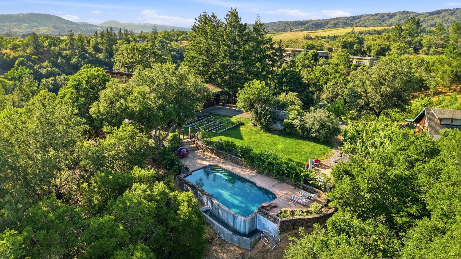 Aerial view of a lush green backyard with a swimming pool surrounded by trees and rolling hills in the background.