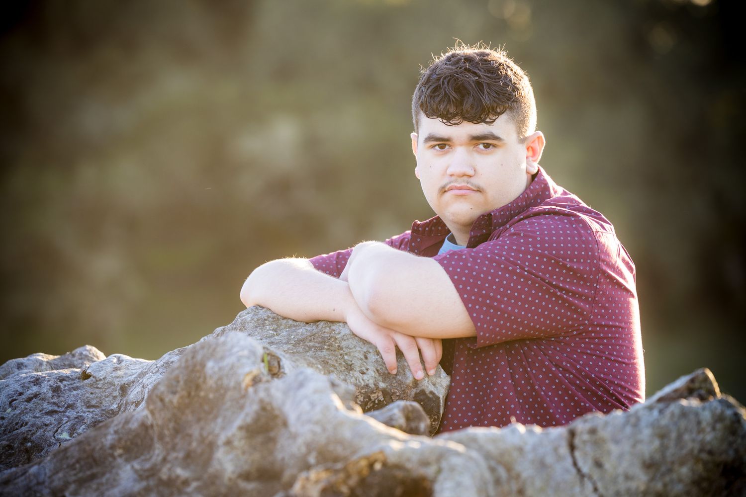 Senior Photos - Portrait of young person sitting on rocky outcrop wearing burgundy shirt at sunset.