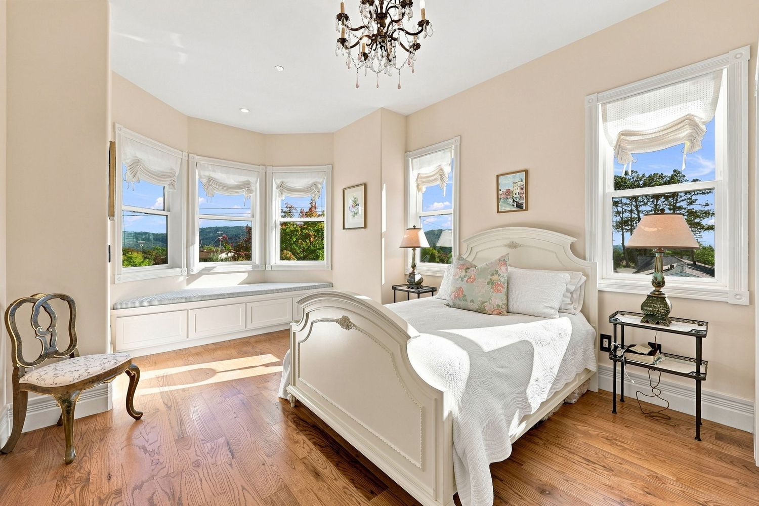 Elegant bedroom with white French bed, hardwood floors, bay windows with scenic views, and a crystal chandelier.