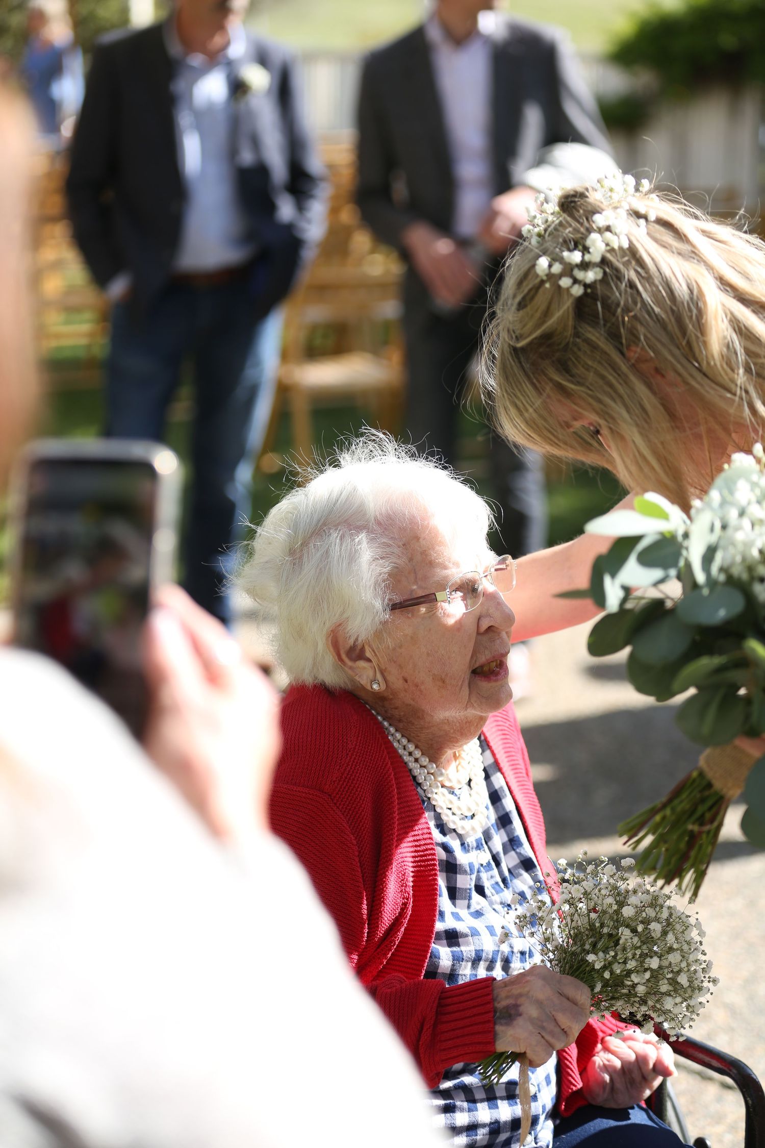 An elderly person in a red cardigan and glasses sits outdoors at a gathering with flowers and guests in the background.