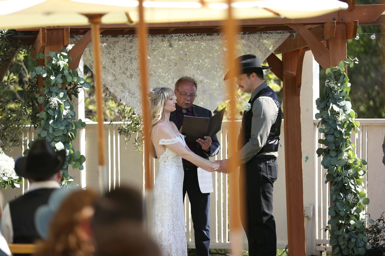 Wedding ceremony takes place outdoors under a wooden gazebo decorated with greenery and white umbrellas.