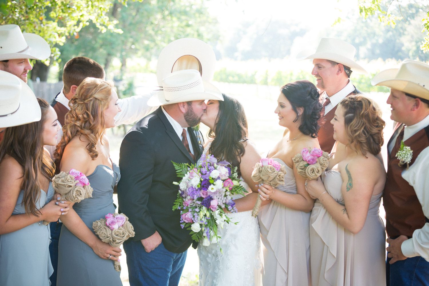 Wedding party in light-colored attire gathers outdoors in sunlit garden setting with purple bouquets.