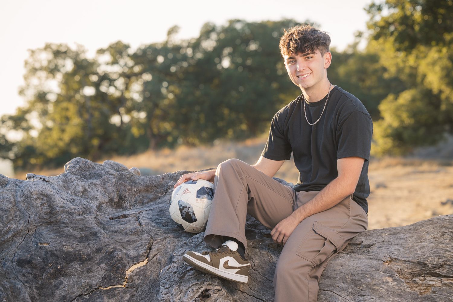 Individual sitting on rock with soccer ball wearing casual attire in outdoor setting.