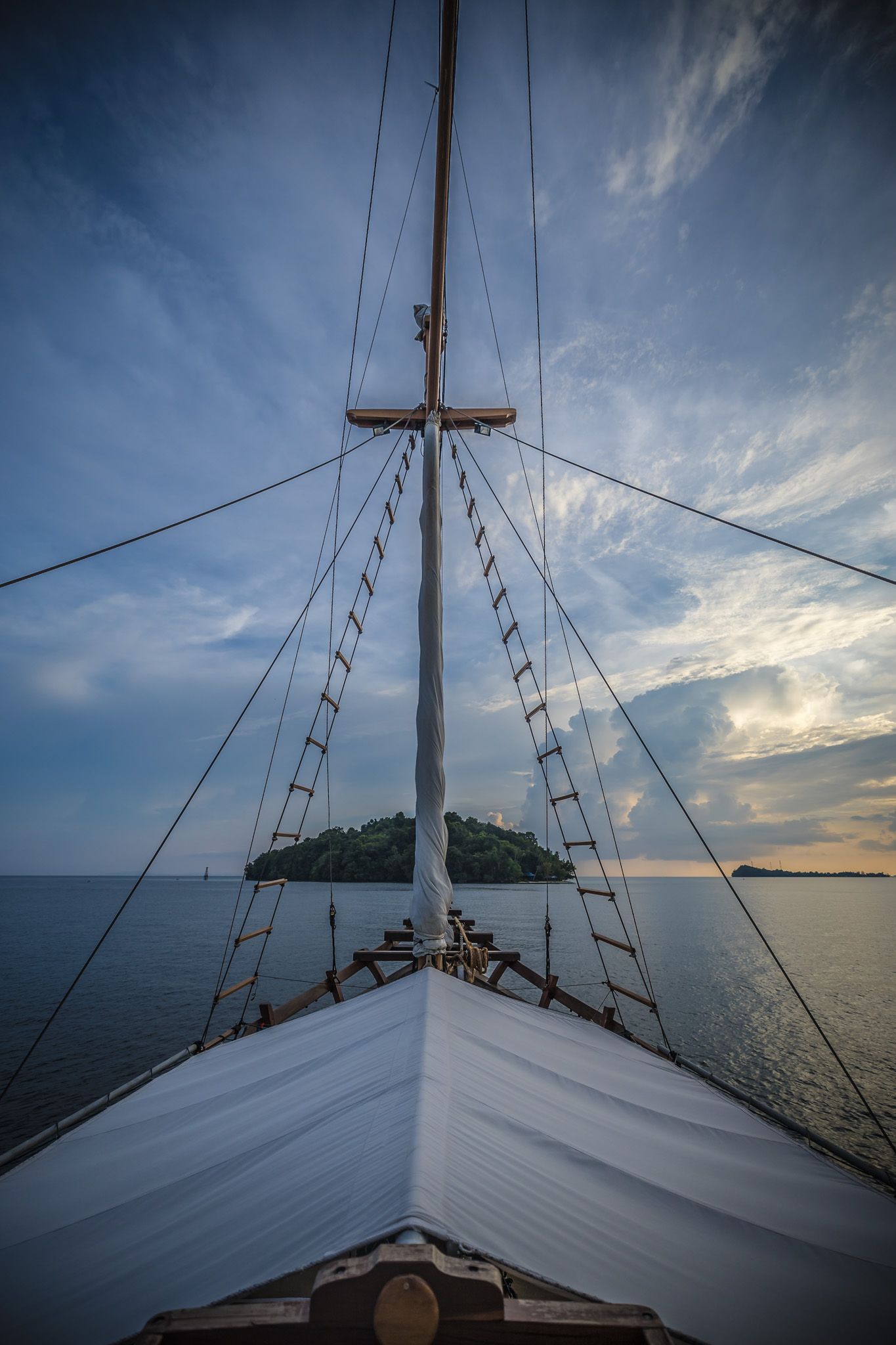 View from ship's bow showing white sails and masts silhouetted against sunset sky.