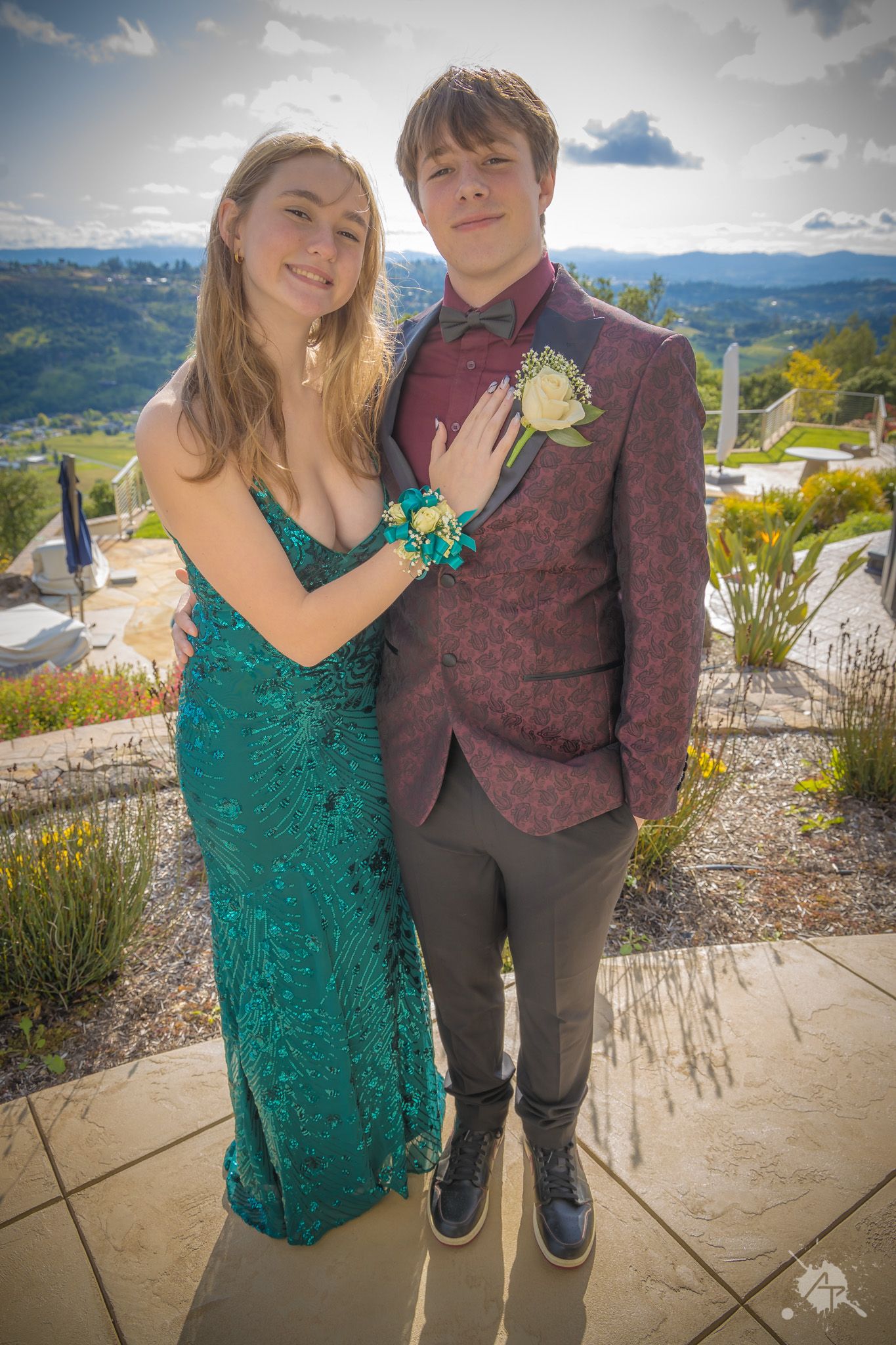 A couple in formal wear poses at a scenic overlook, with her in emerald green and him in burgundy.