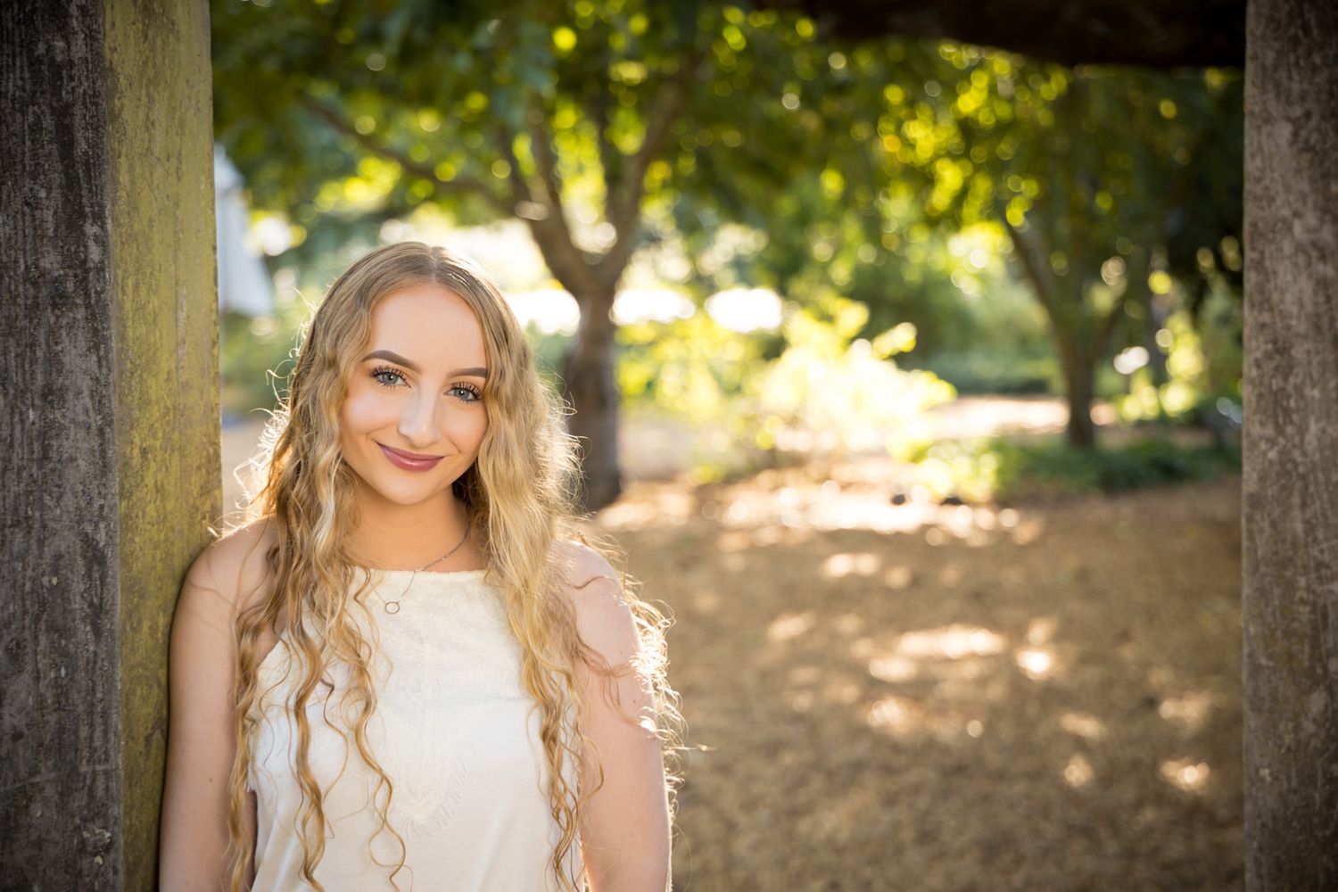 A portrait in a sunlit park shows a white summery dress and long blonde curls against a soft nature backdrop.
