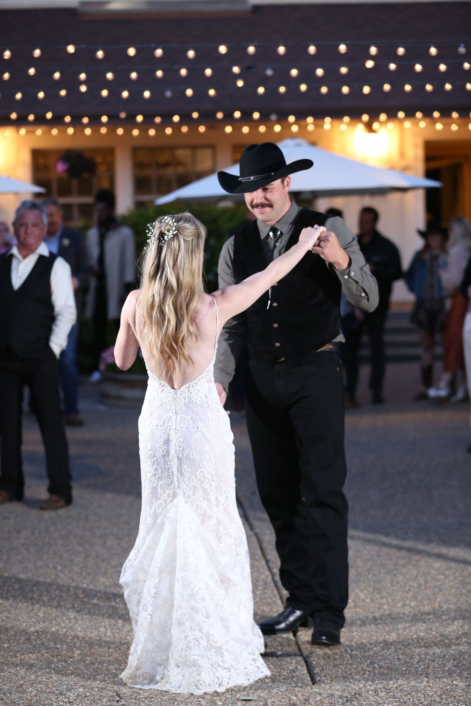 A bride in a white lace gown dances with someone wearing a cowboy hat and black attire under twinkle lights.
