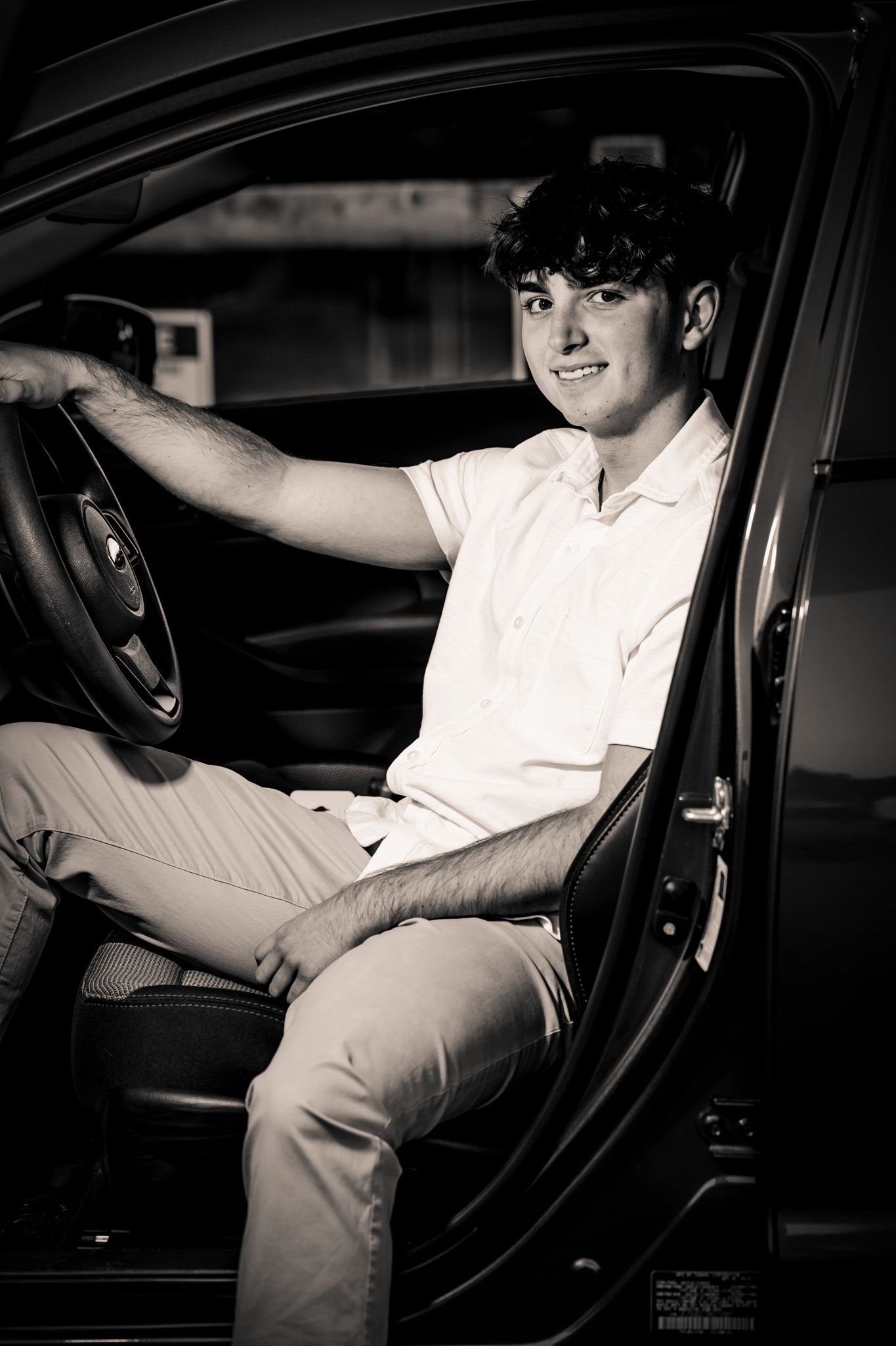 Individual sitting in driver's seat of car wearing white polo in black and white portrait.