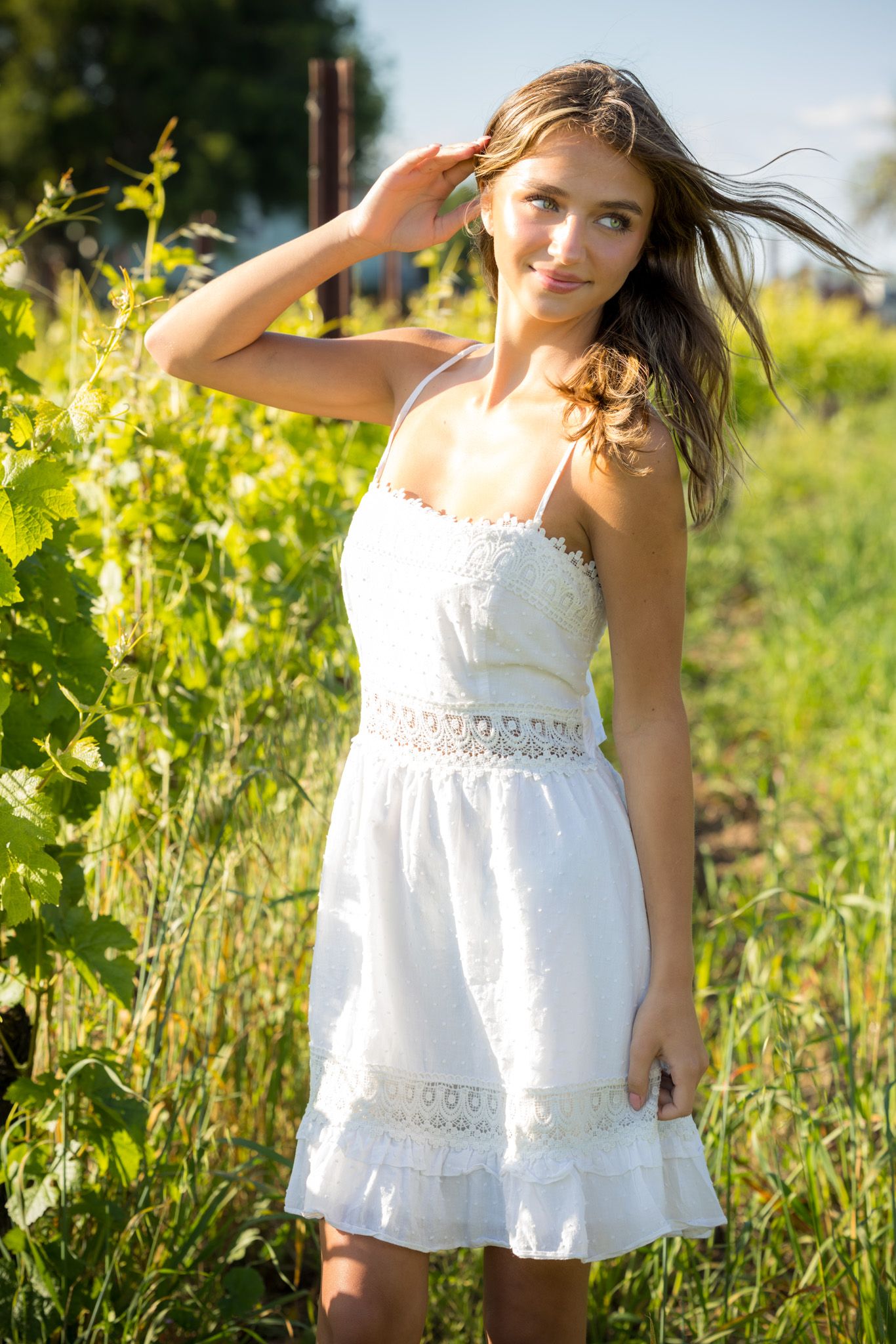 Young woman in white sundress standing among green vineyard plants on a sunny day.