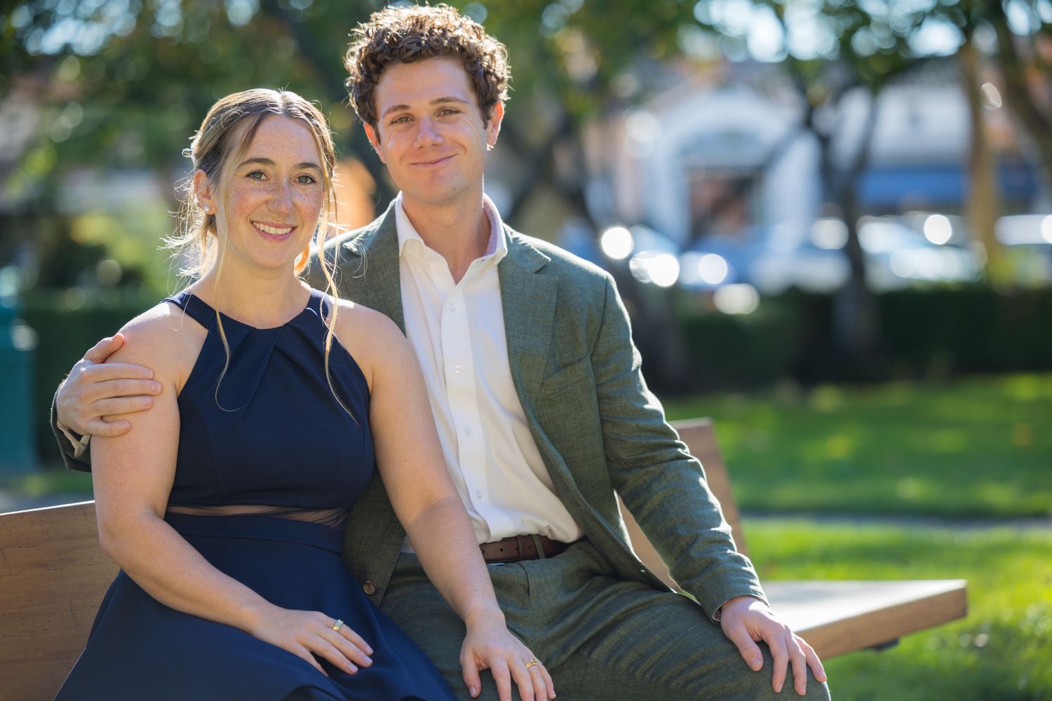 Smiling couple sitting together on a bench in a sunny park, woman in navy dress and man in green blazer.