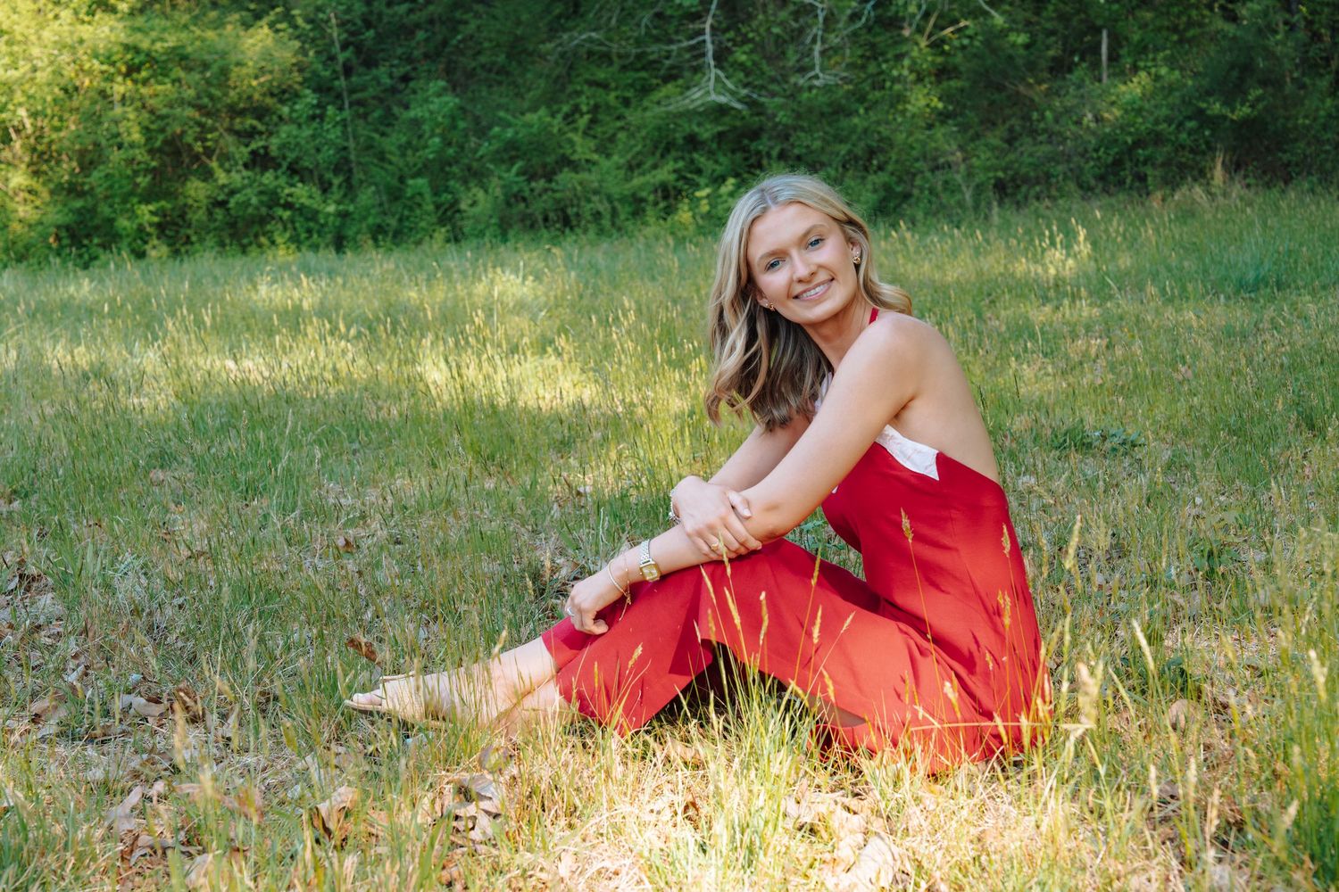 Smiling young blonde woman in red outfit sitting in a sunny green meadow surrounded by trees.