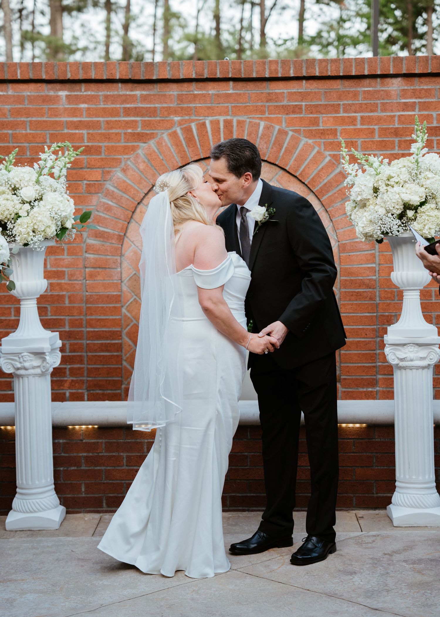 Bride and groom share first kiss at outdoor brick arch altar adorned with white floral arrangements on white columns.