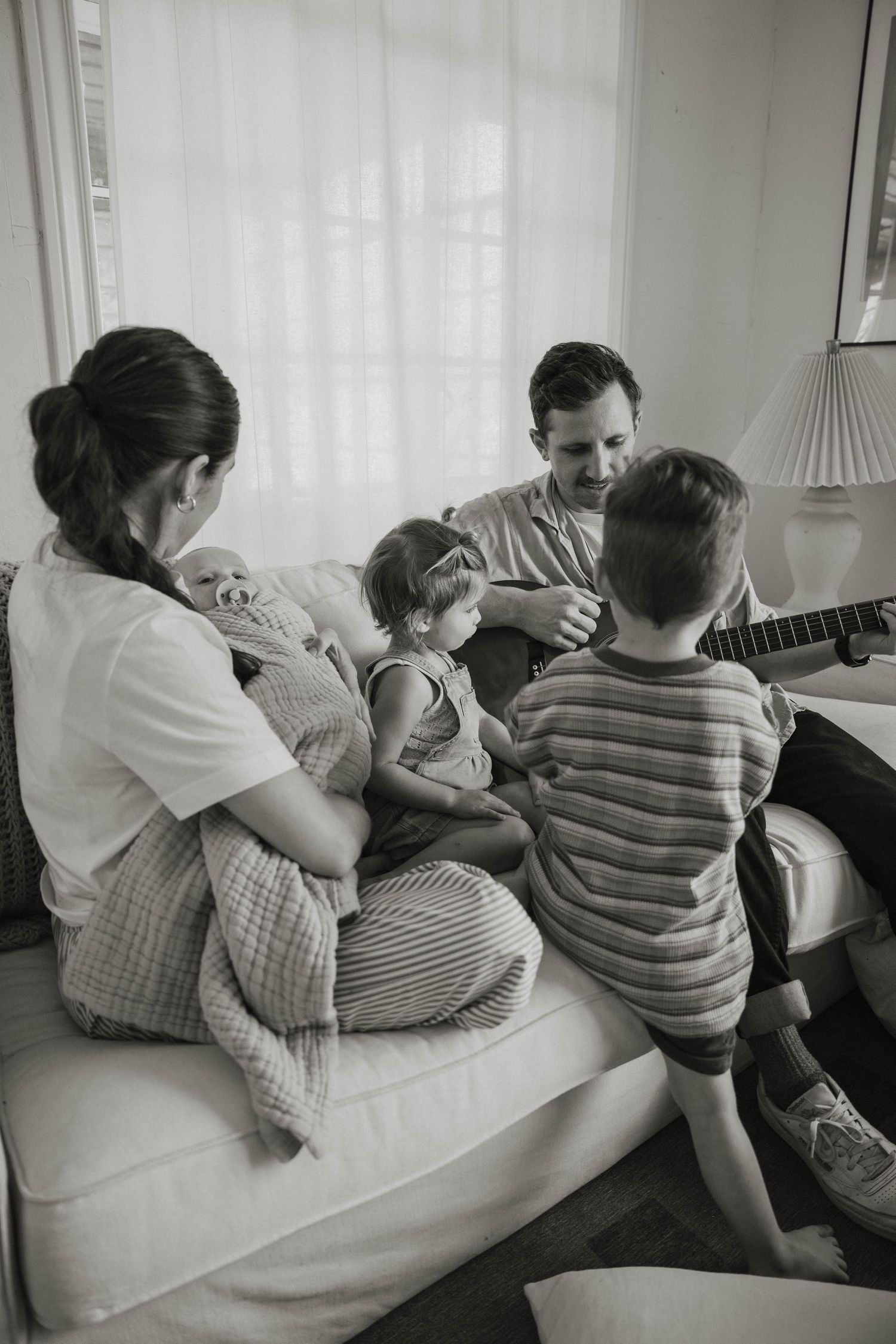 Family gathered on couch sharing musical moment with acoustic guitar and children in black and white photo.
