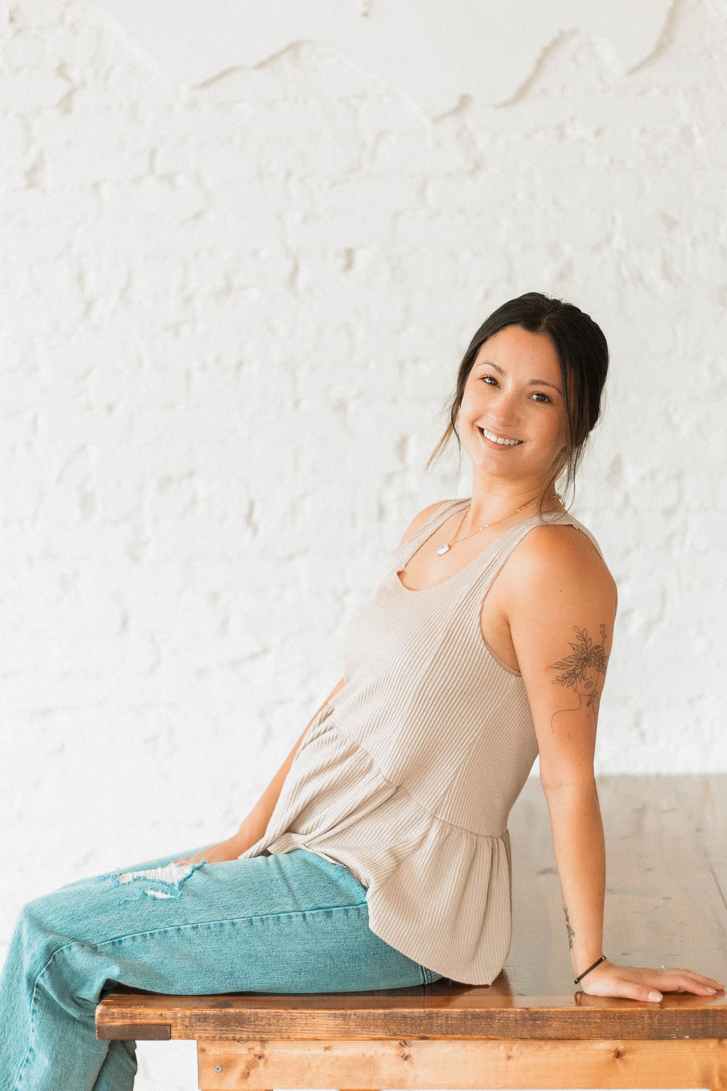 Woman in beige top and teal pants sits casually on wooden bench against white textured wall.