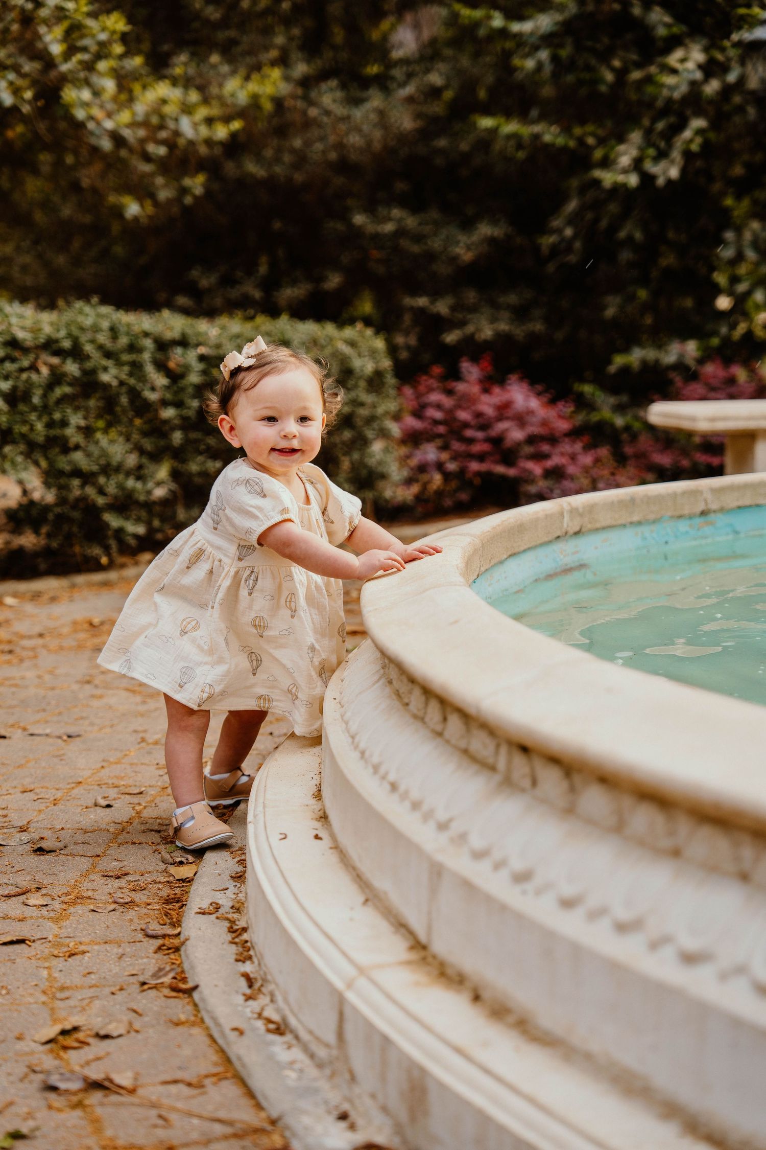 A small child in a white dress plays near a curved stone fountain on fallen autumn leaves.