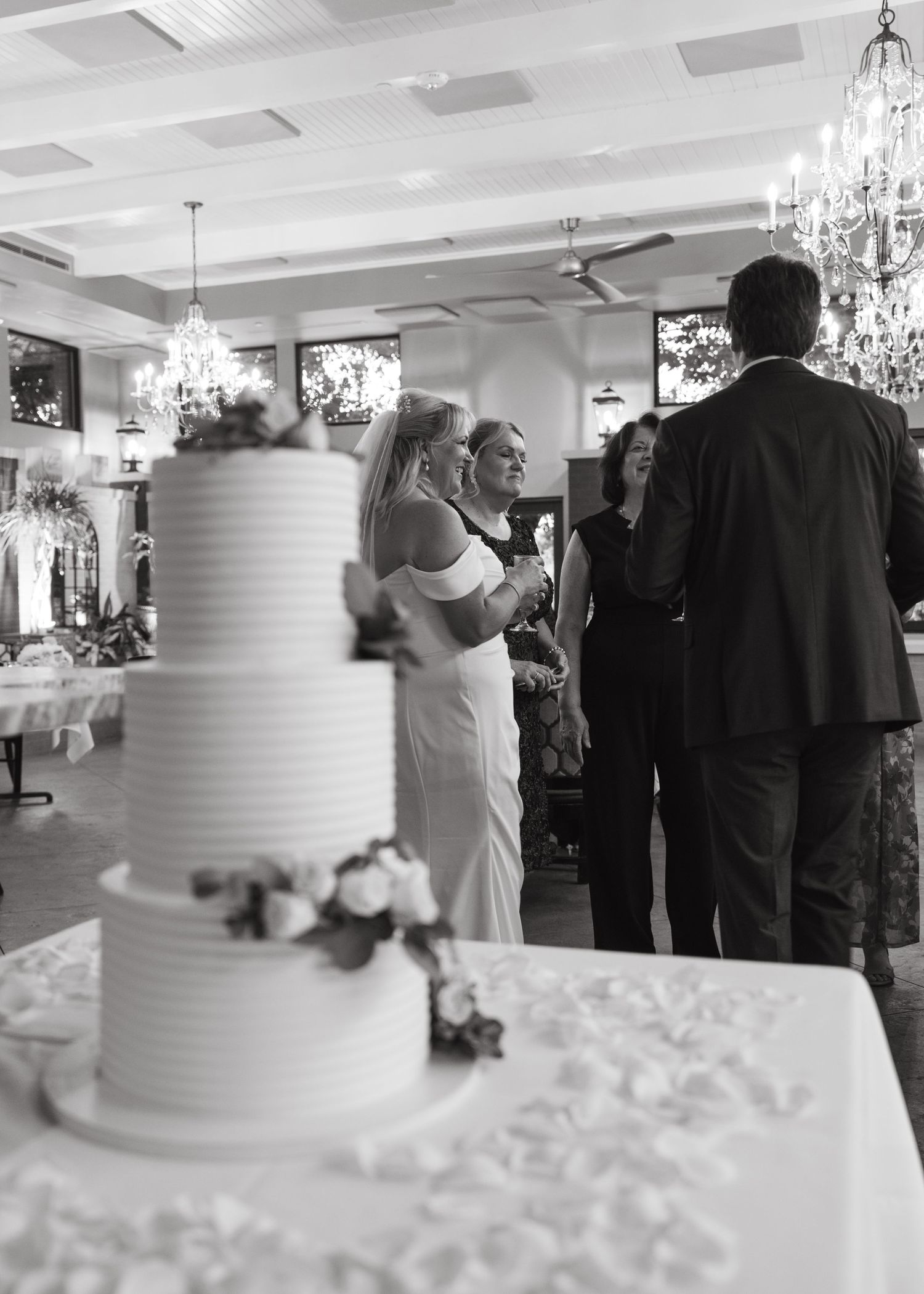 Black and white wedding reception photo featuring elegant tiered cake with floral decor and guests in formal attire.