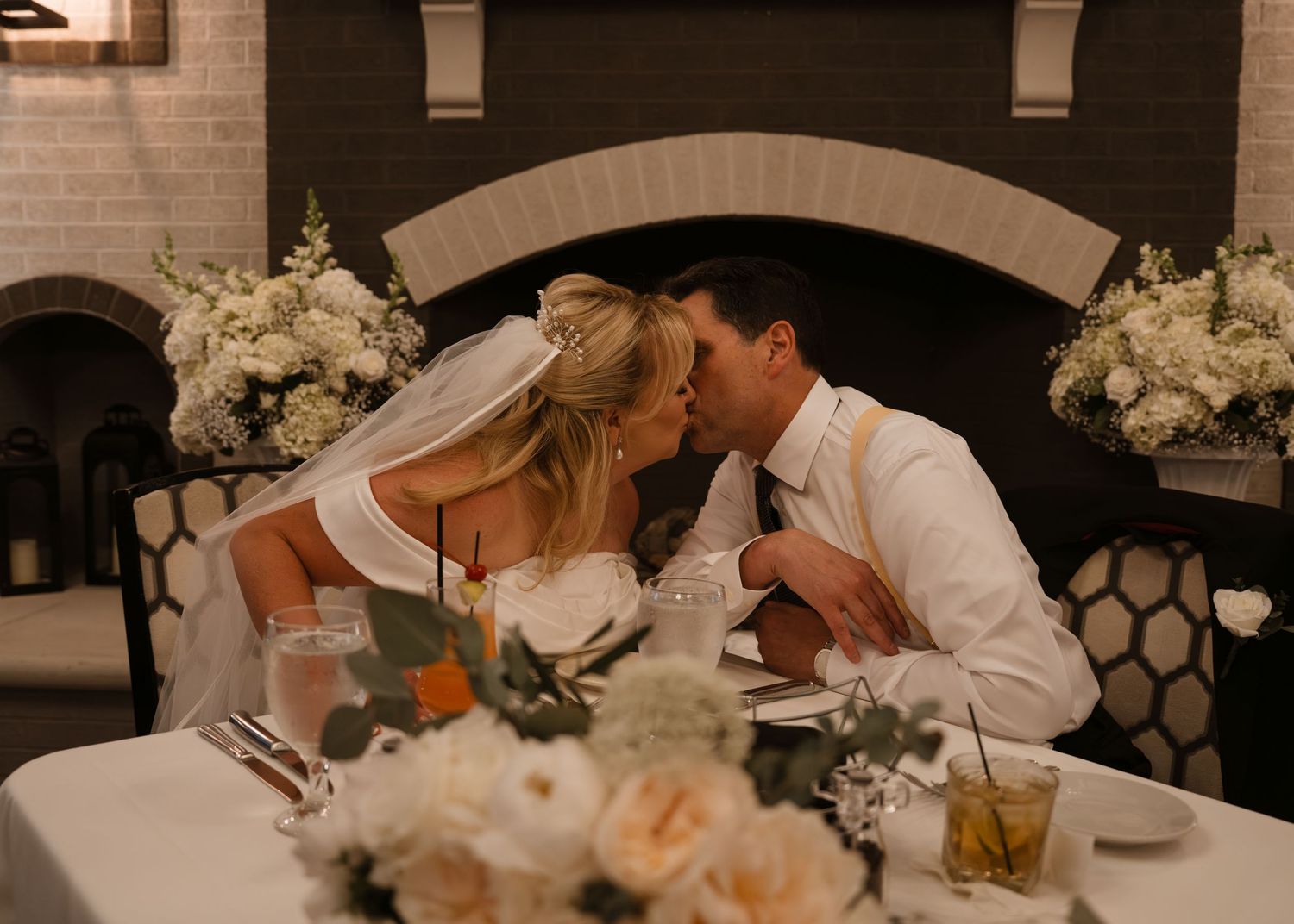 Bride and groom sharing a kiss at their sweetheart table with elegant white floral arrangements at their wedding reception.