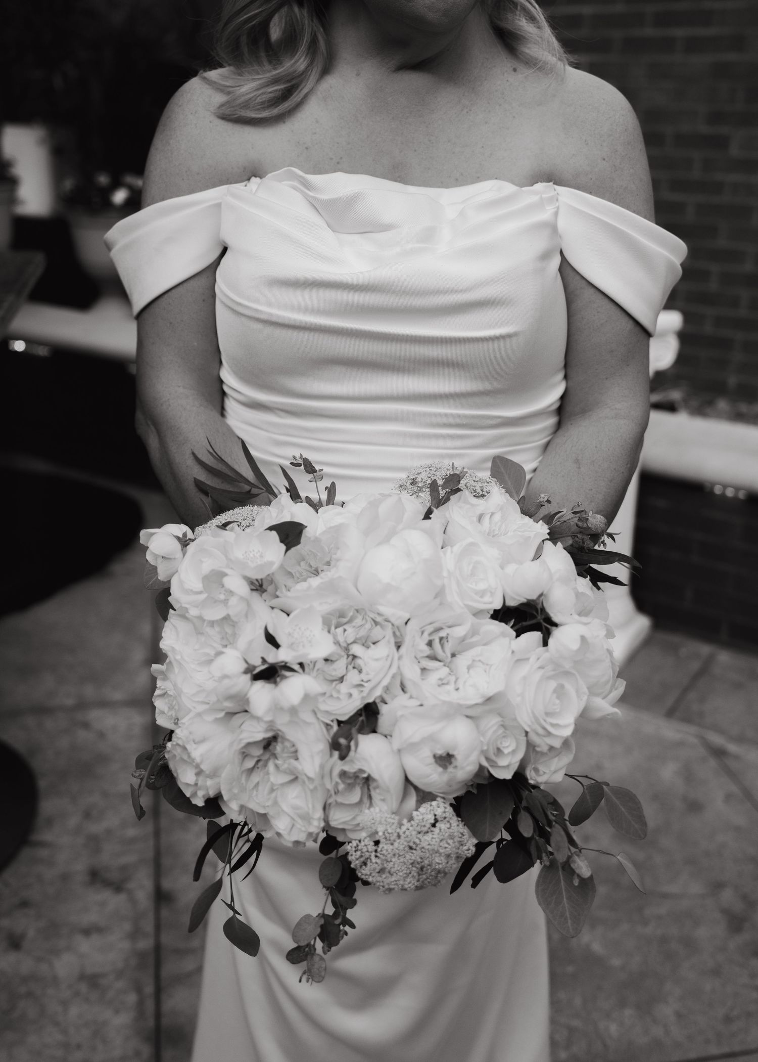Bride in off-shoulder white gown holding large bouquet of white roses and peonies, black and white wedding photo.