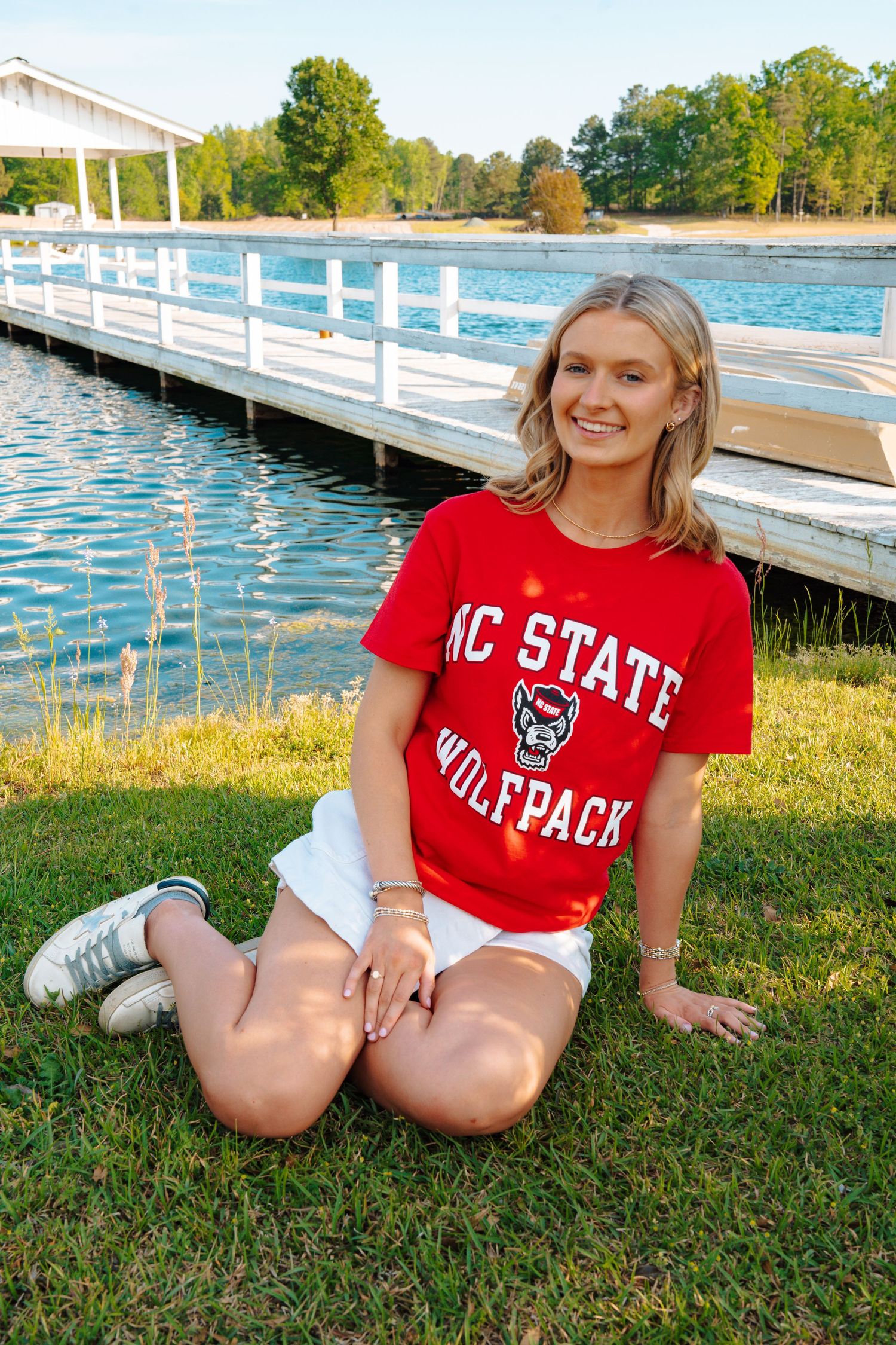 Smiling blonde woman in NC State Wolfpack red shirt sitting on grass by a lake with a wooden dock.