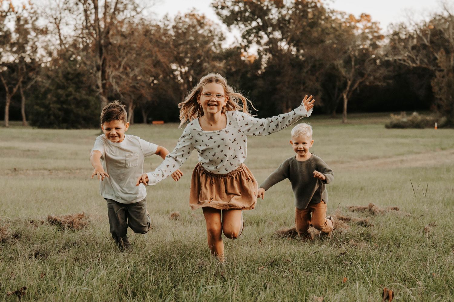 Three happy children running through a green meadow in autumn, laughing and holding hands outdoors.