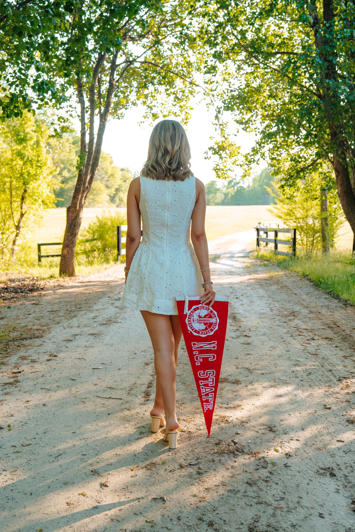 Blonde woman in white dress walking down a sandy path holding a red pennant flag surrounded by green trees.