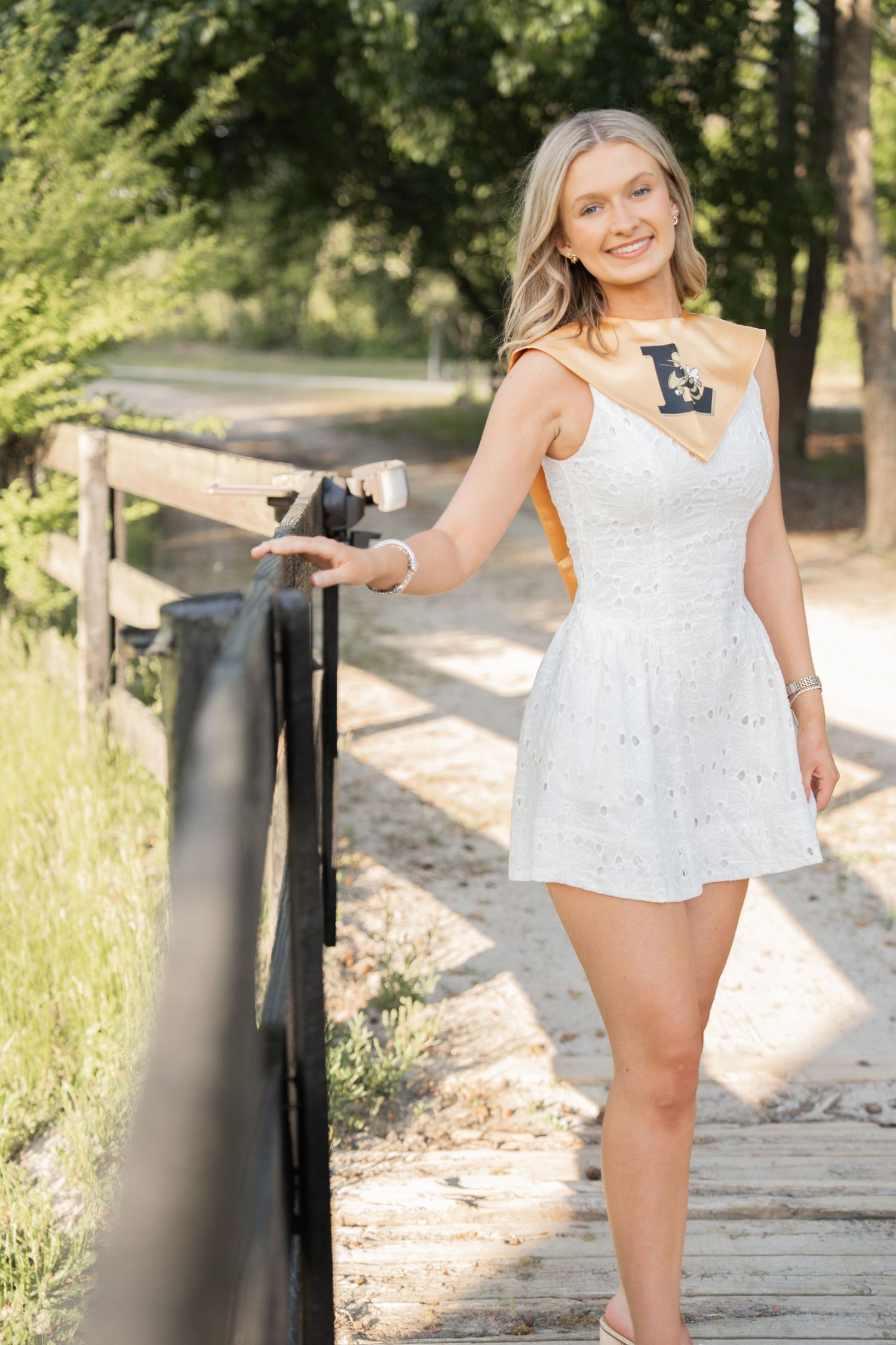 Smiling blonde woman in white dress holding railing on wooden bridge surrounded by green trees outdoors.