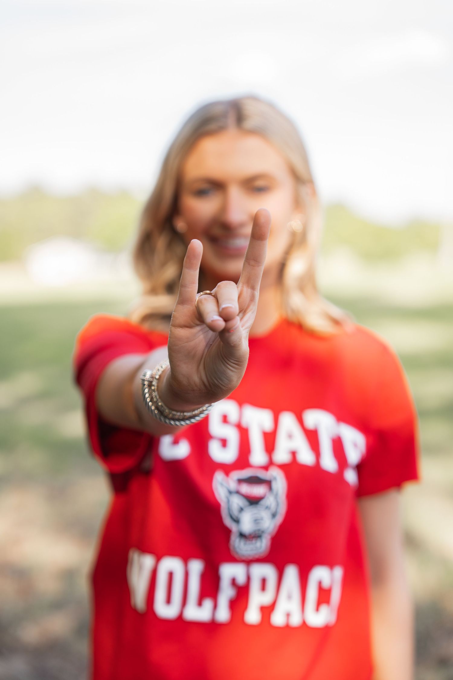 A smiling blonde woman in an NC State Wolfpack red shirt makes the wolf sign hand gesture toward the camera.