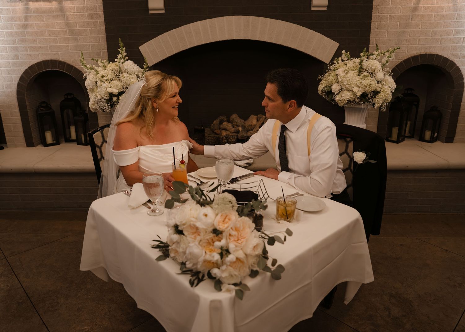Bride and groom share an intimate moment at their elegant sweetheart table beside a white brick fireplace.
