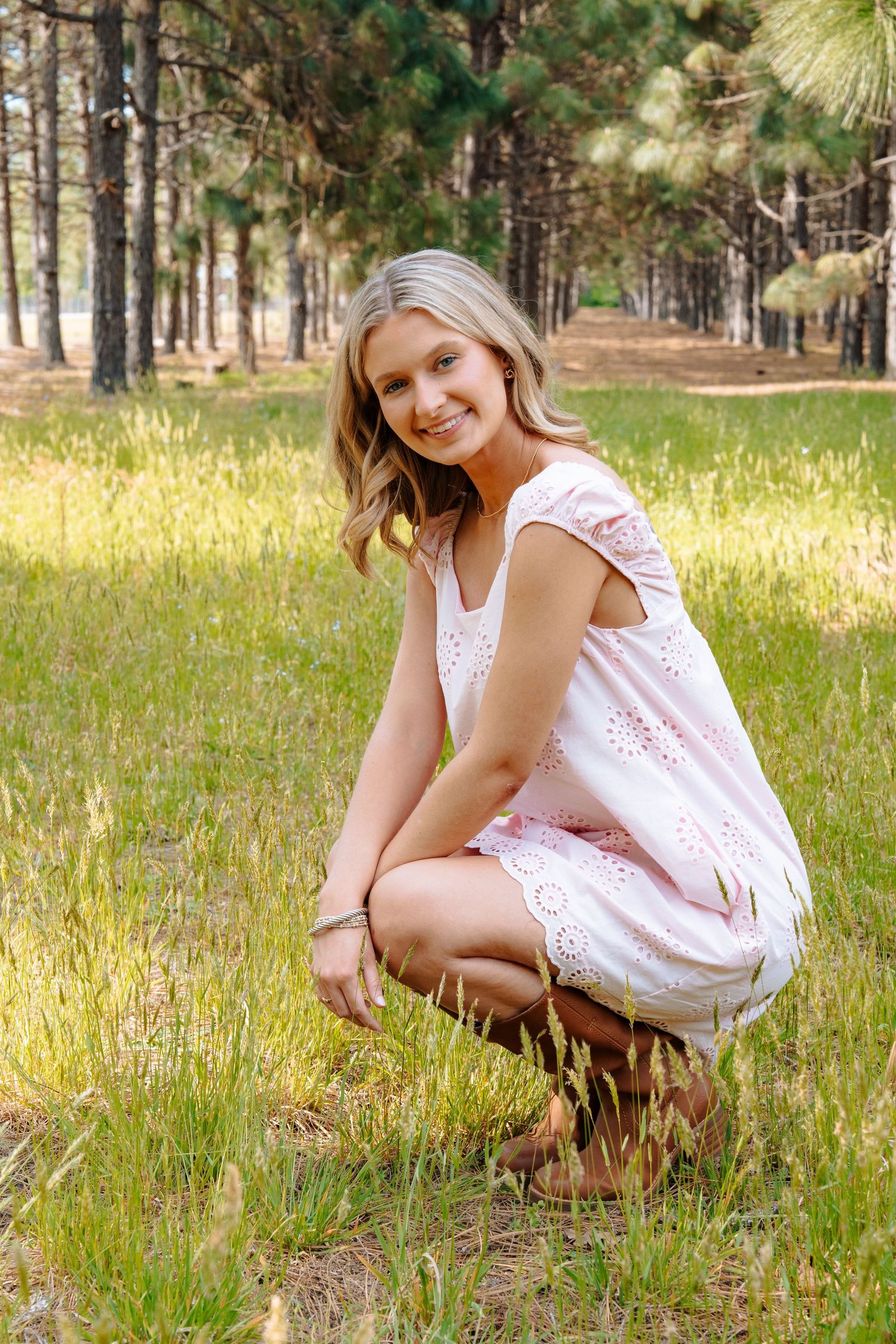 Young blonde woman in pink eyelet dress crouching in a sunlit forest clearing, smiling at the camera.