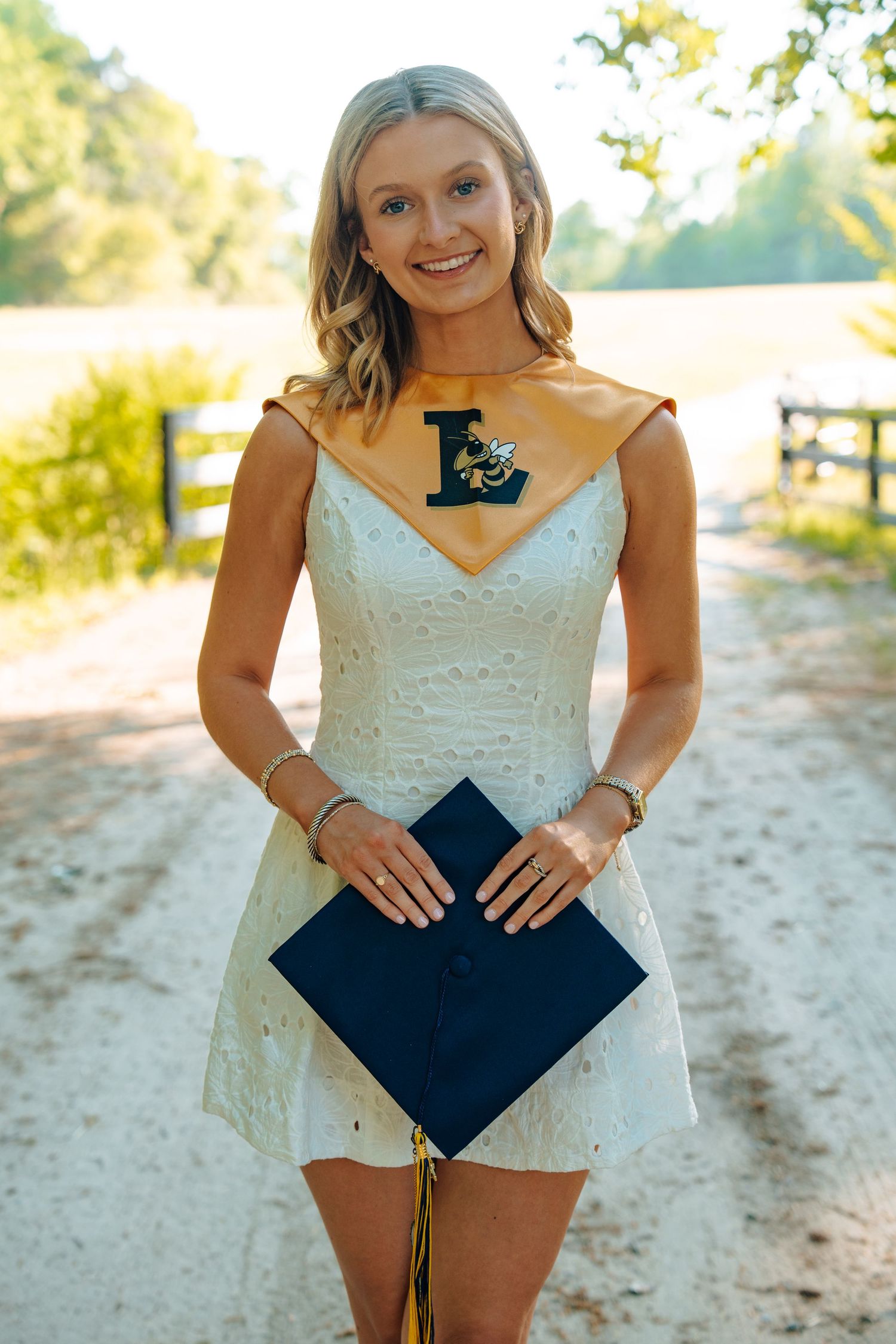 Smiling blonde graduate in white dress holds navy cap, wearing gold graduation stole with letter L on a sunny gravel path.