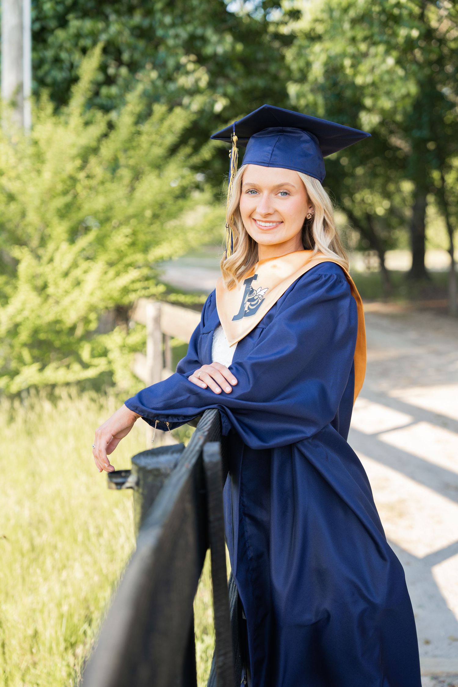 Smiling female graduate in navy blue cap and gown posing outdoors by a fence with green trees in background.