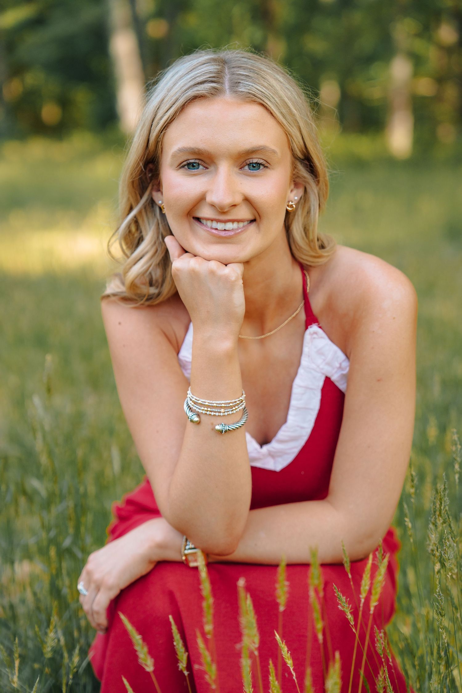 Smiling blonde woman in red dress sitting in green grass field with hand on chin.