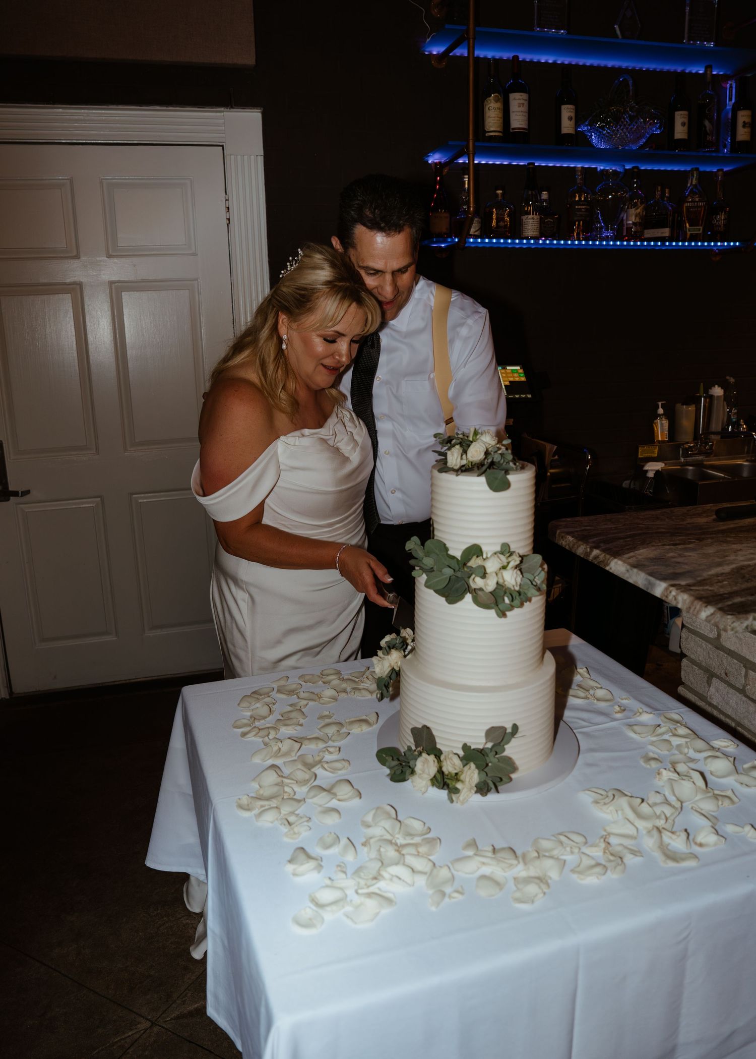 Bride and groom cutting a three-tier white wedding cake decorated with greenery on a table with rose petals.
