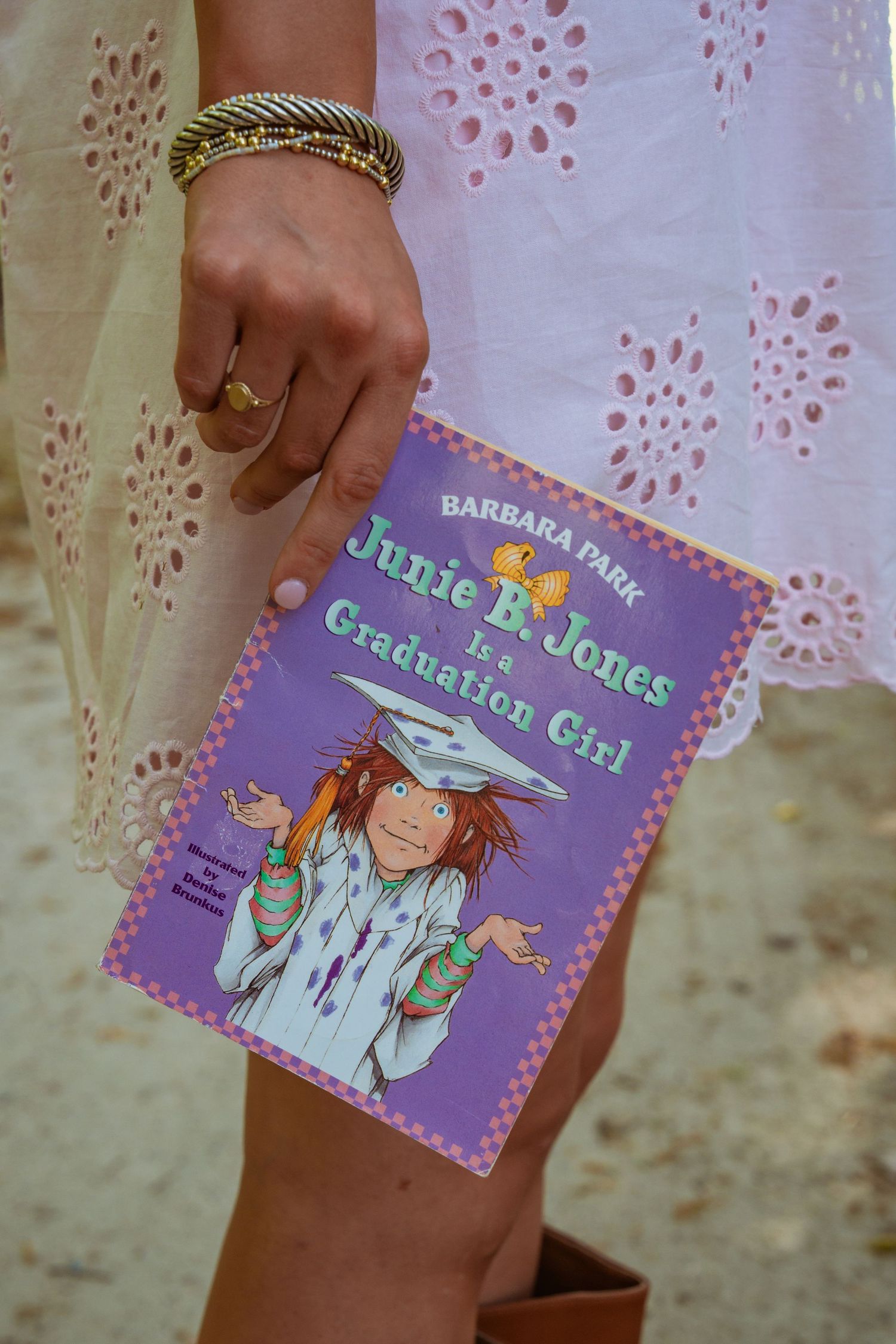 A hand holding Junie B. Jones Is a Graduation Girl book by Barbara Park, wearing gold bangles and a ring.