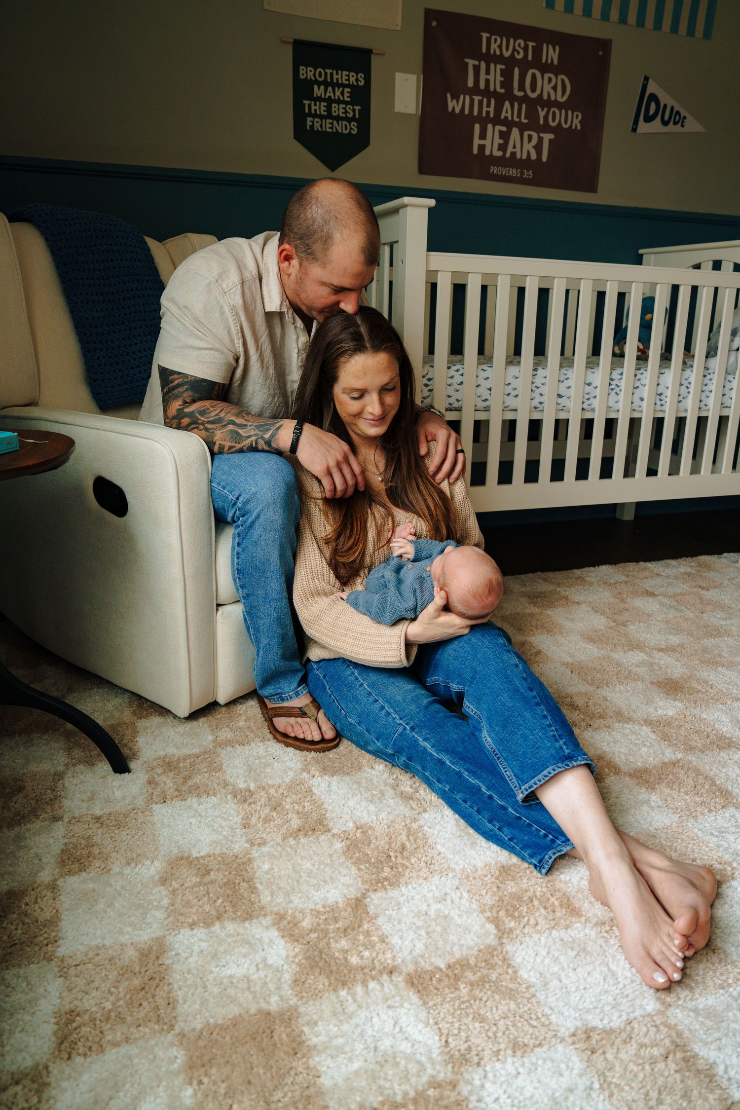 A family sits together on the floor of a nursery with a white crib and religious wall decor in the background.