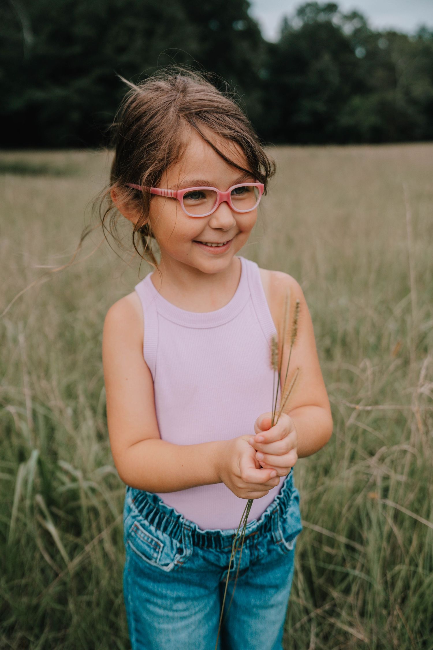Child in pink tank top and jeans holds wheat stalk while playing in tall grass field on sunny day.