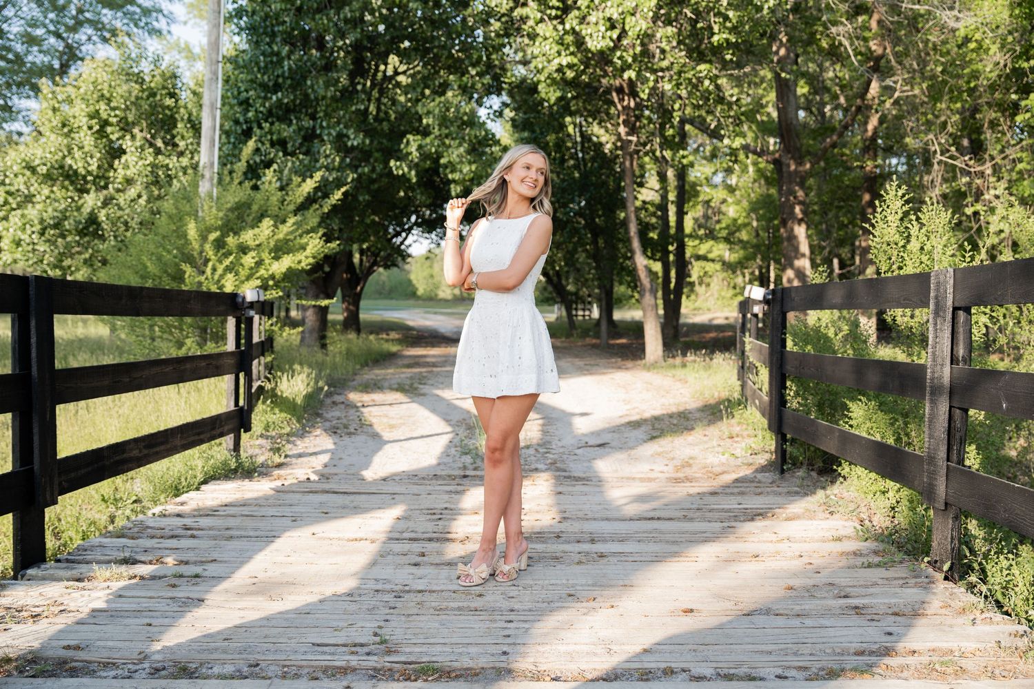 Young woman in white dress standing on a wooden bridge surrounded by lush green trees on a sunny day.
