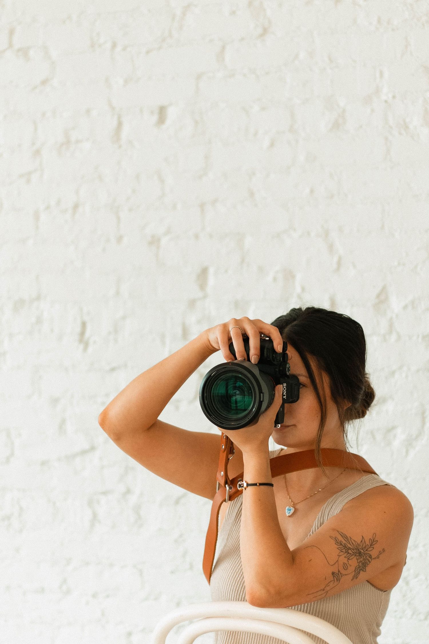 A photographer wearing a white tank top takes a photo with a professional camera against a white textured wall.