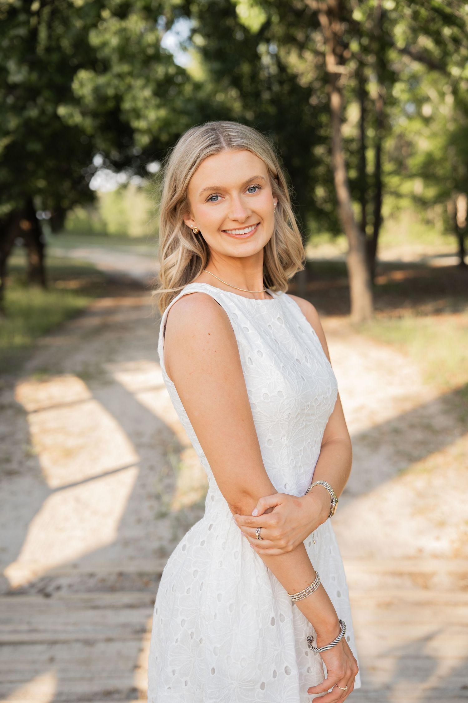 Smiling blonde woman in white sleeveless dress standing on a sunlit tree-lined path outdoors.