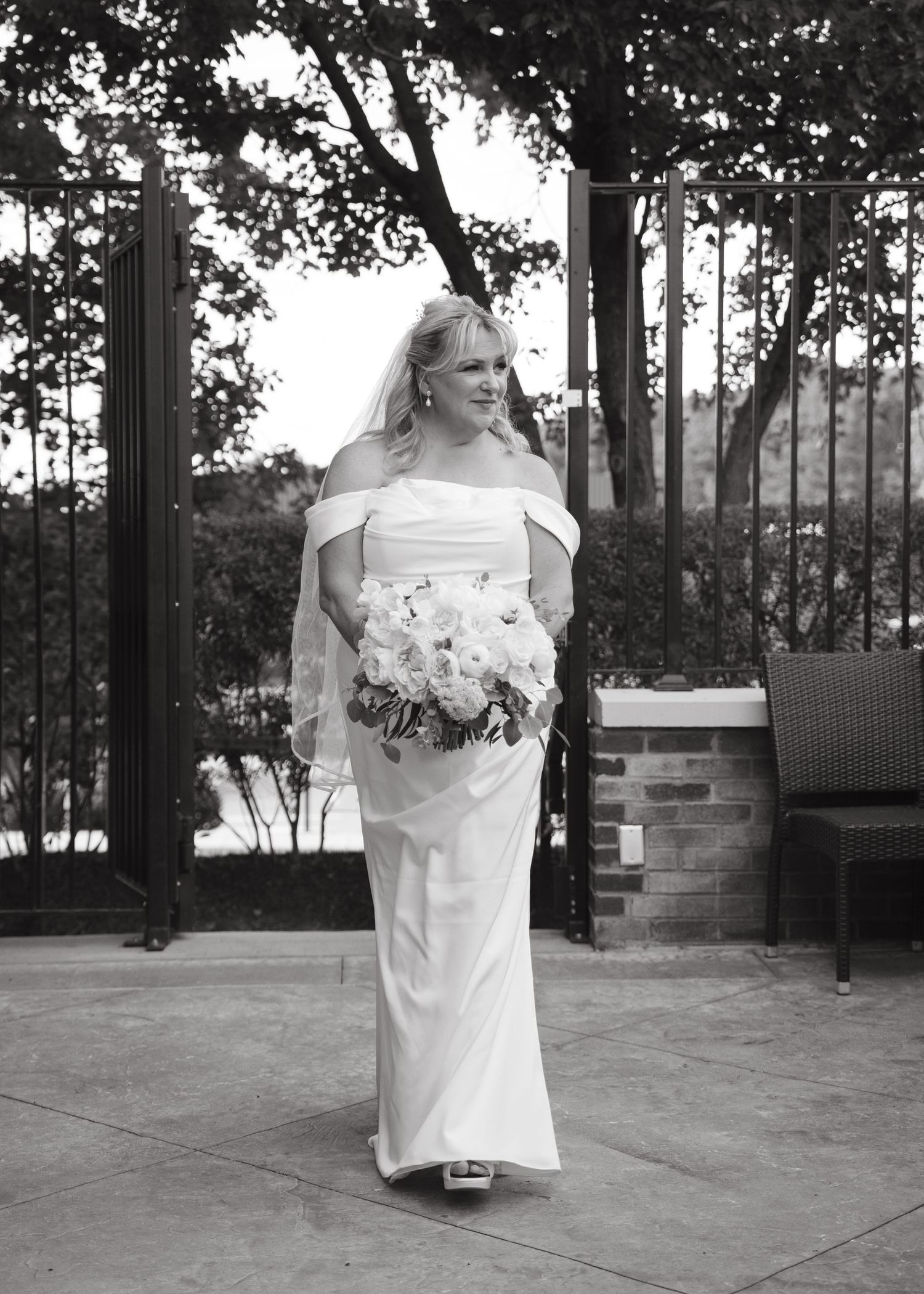 Bride in off-shoulder white gown holding bouquet walks on patio in black and white wedding photo.