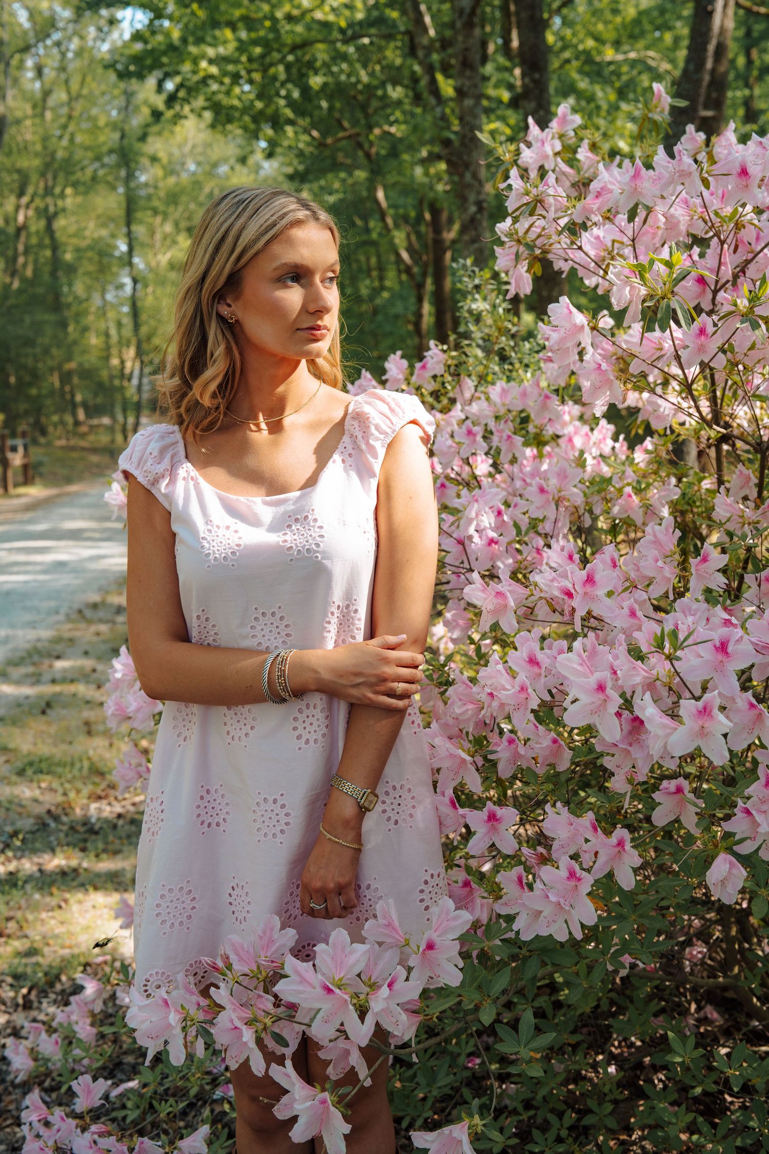 Young blonde woman in white eyelet dress standing beside pink azalea blooms outdoors.