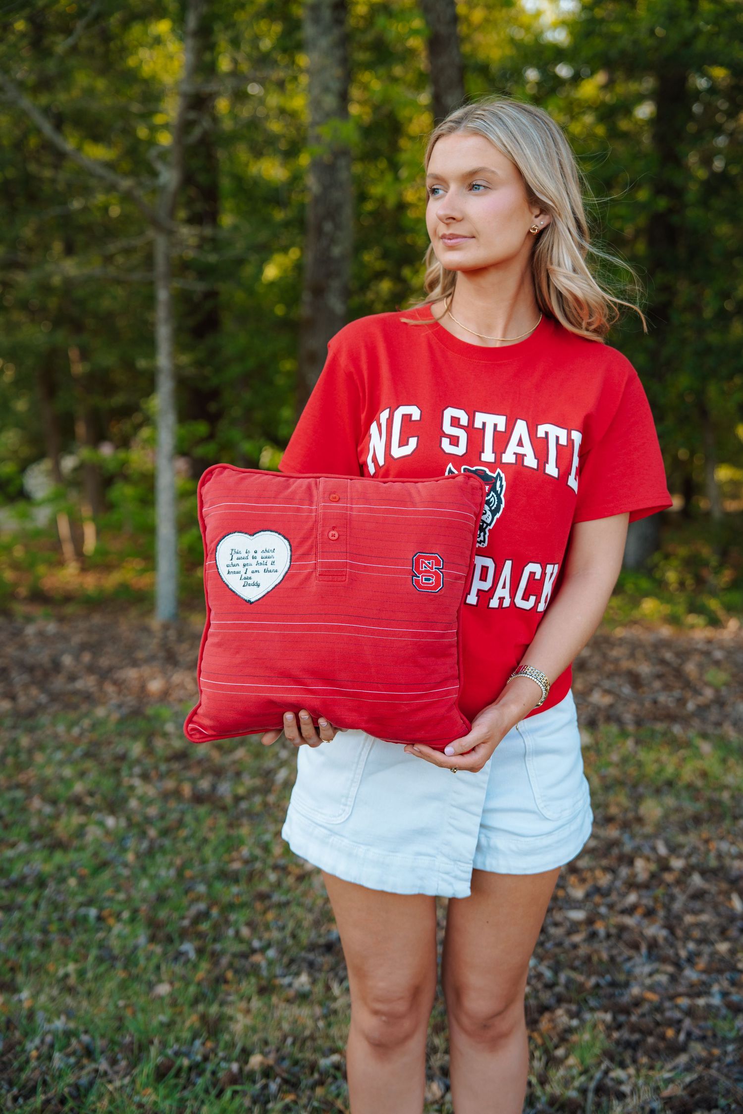 Young woman in red NC State Wolfpack t-shirt holding a red decorative pillow outdoors among trees.
