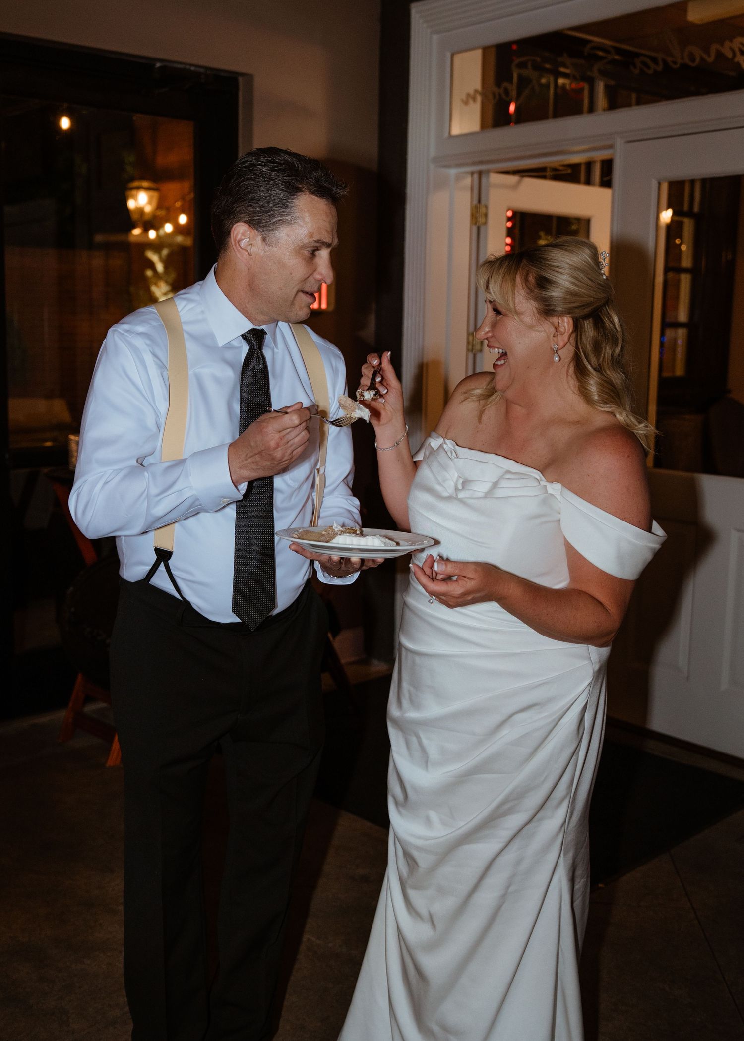 Bride in white off-shoulder gown and groom in suspenders share wedding cake during indoor reception.