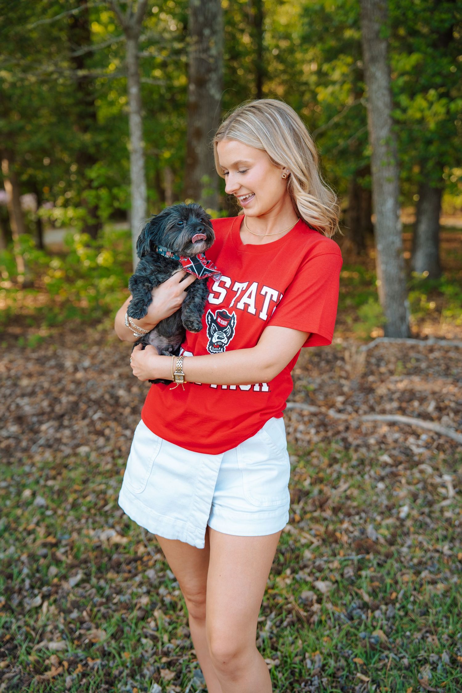Blonde woman in red NC State shirt holding a small black dog outdoors near trees.