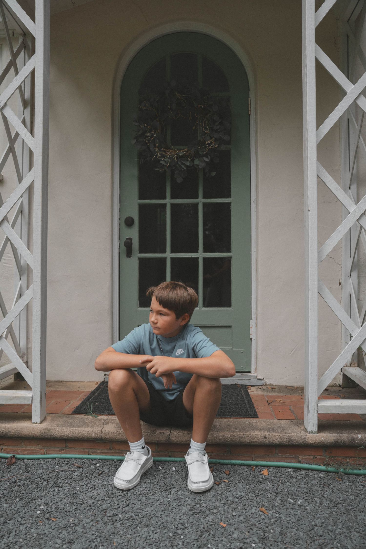 A child sits on a porch with white lattice work and green doors in a contemplative pose.