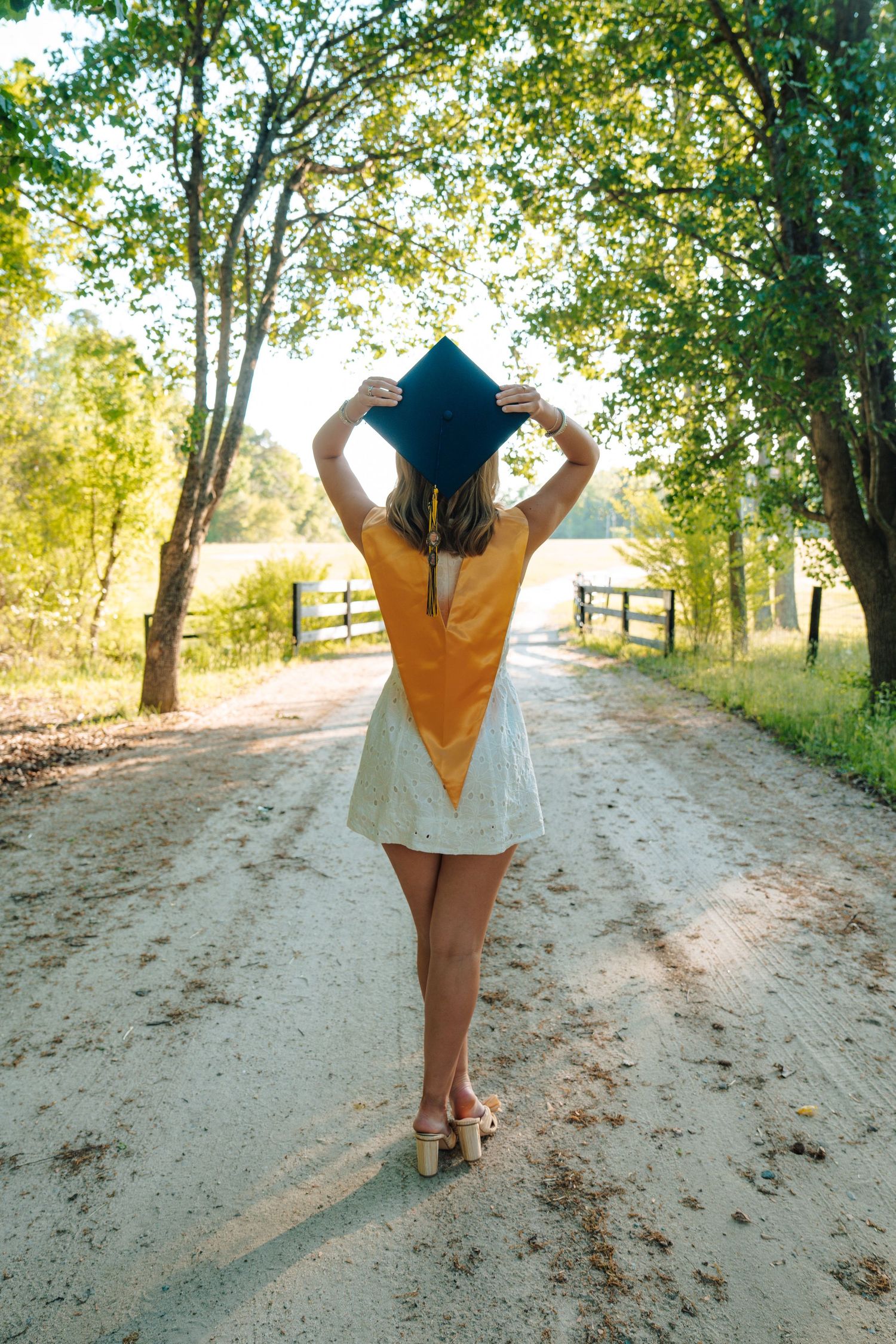 A female graduate stands on a rural dirt road holding her cap, wearing a yellow graduation stole over a white dress.